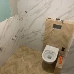 Wnętrze łazienki w trakcie remontu: częściowo położone marmurowe płytki ścienne, podłoga wyłożona płytkami w jodełkę, zamontowana podtynkowa spłuczka WC z widocznymi przewodami, biała miska...