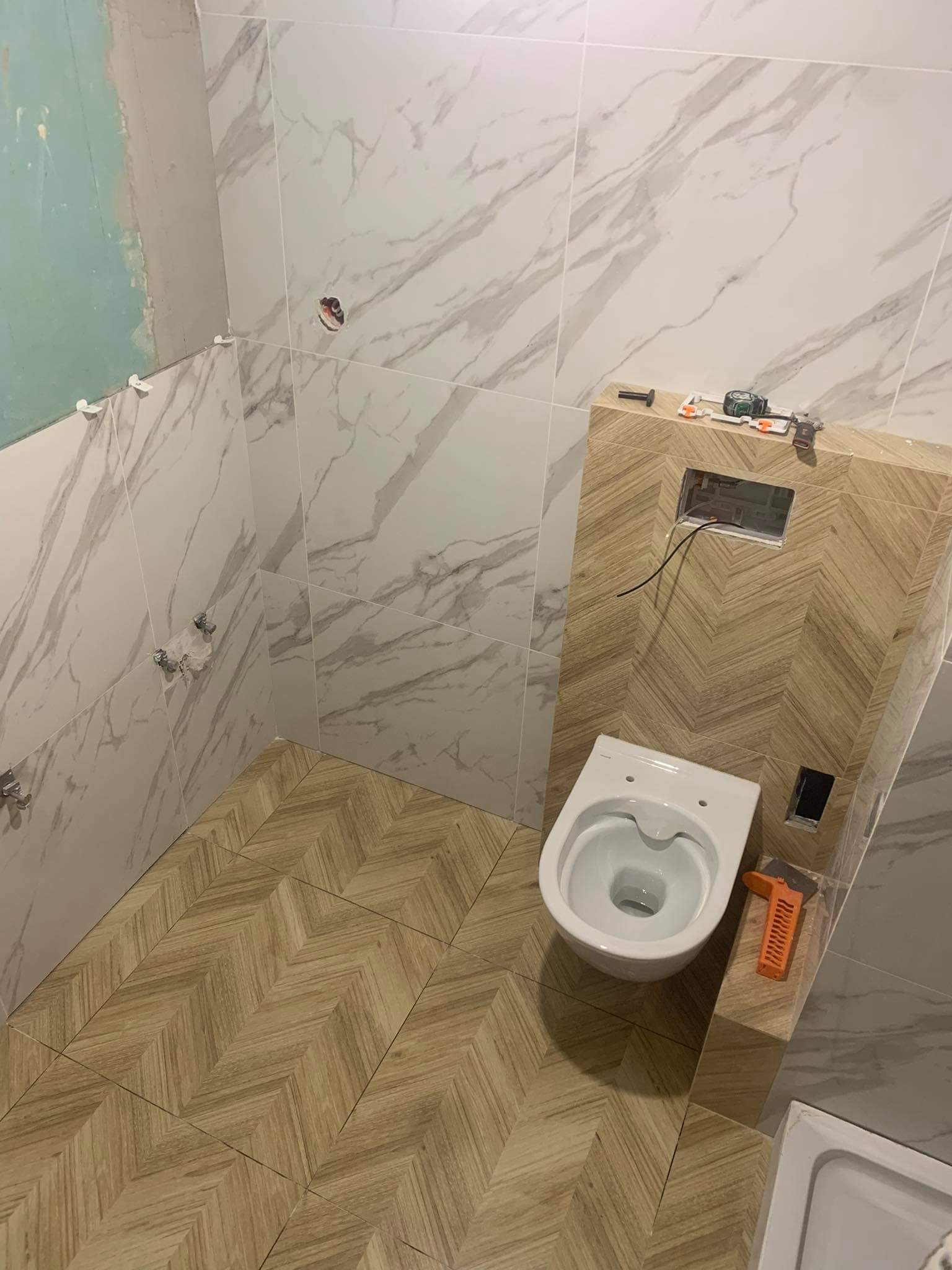 Wnętrze łazienki w trakcie remontu: częściowo położone marmurowe płytki ścienne, podłoga wyłożona płytkami w jodełkę, zamontowana podtynkowa spłuczka WC z widocznymi przewodami, biała miska...