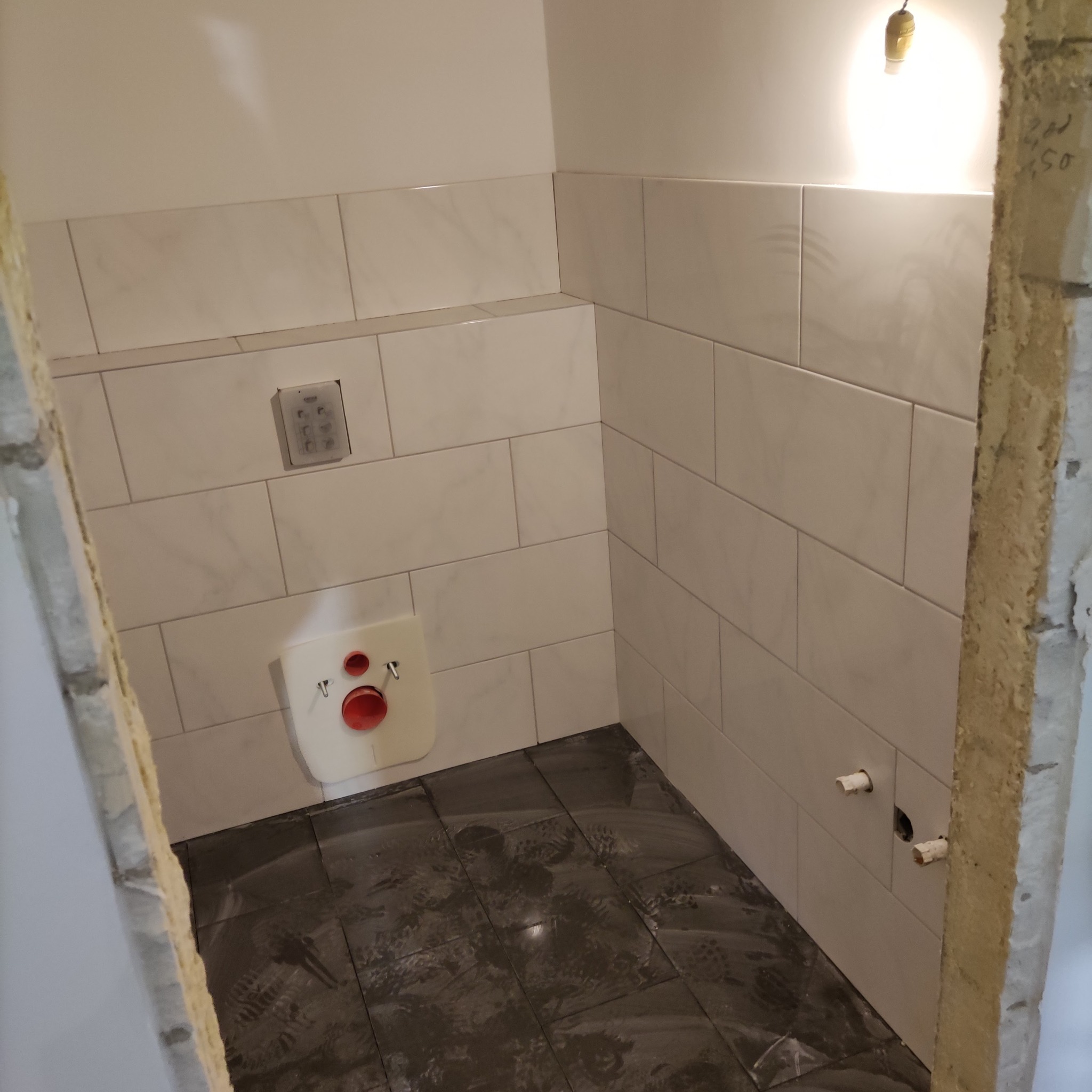 Wnętrze łazienki w trakcie remontu: ułożone białe płytki na ścianach, ciemne płytki na podłodze, widoczna instalacja podtynkowego systemu spłukiwania, wystające rury instalacji wodnej, surowy otwór...