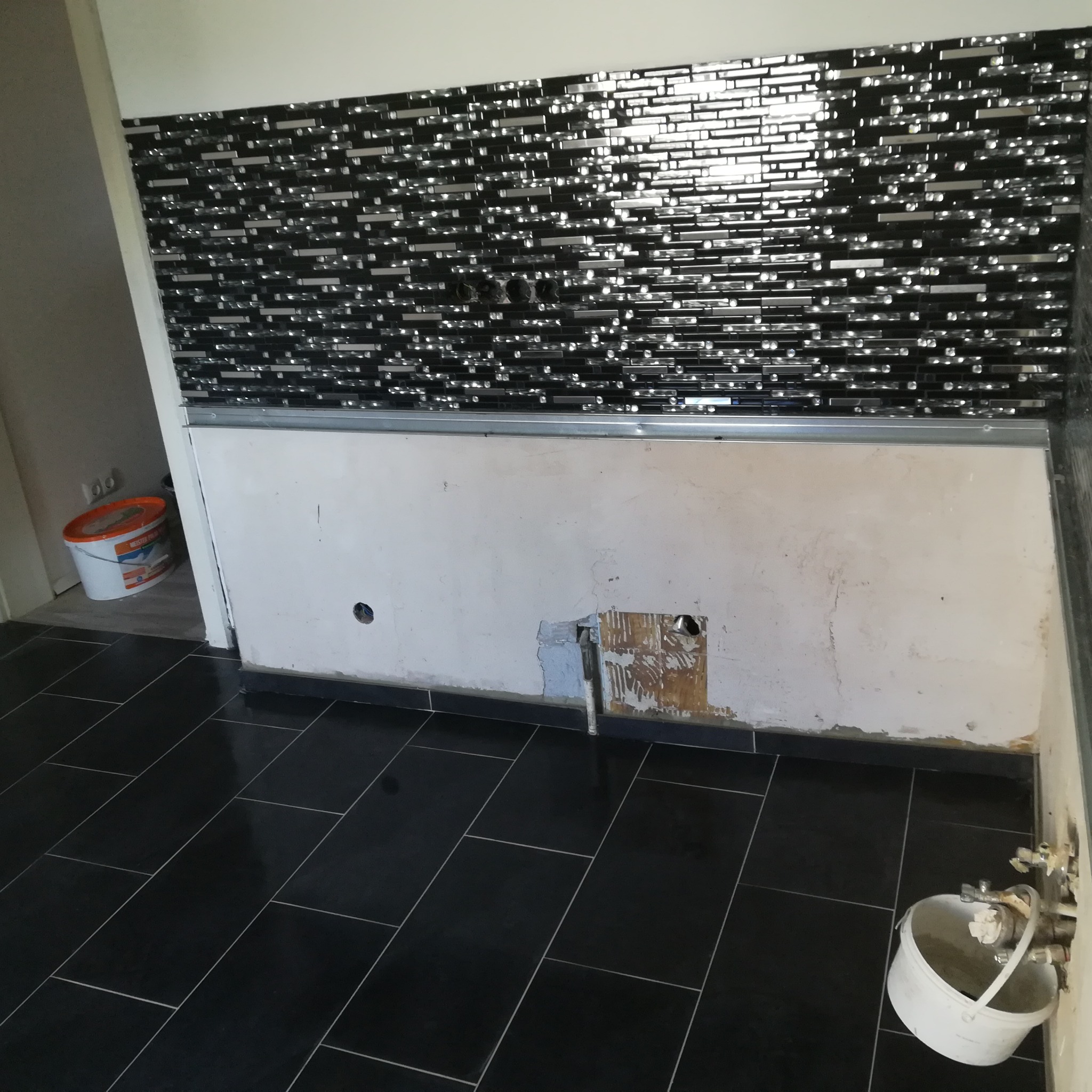 Częściowo wykończona ściana z czarno-srebrną mozaiką w łazience, widoczne przygotowania do instalacji wodnej i elektrycznej, czarna błyszcząca podłoga, ślady prac remontowych.