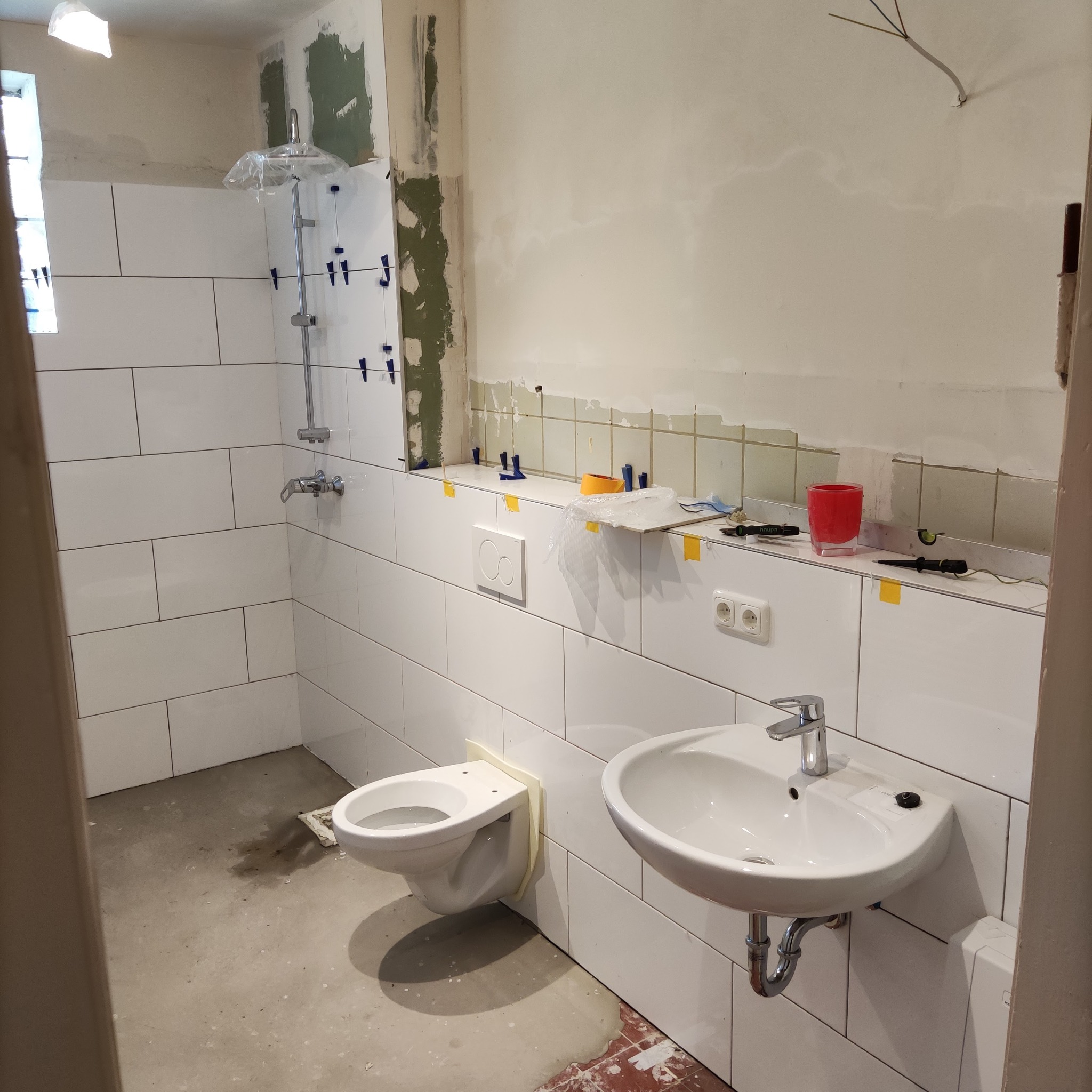 Wnętrze remontowanej łazienki we Włocławku: białe płytki ułożone na ścianach, zamontowany prysznic z deszczownicą, częściowo podłączona toaleta i umywalka, widoczne narzędzia glazurnicze...