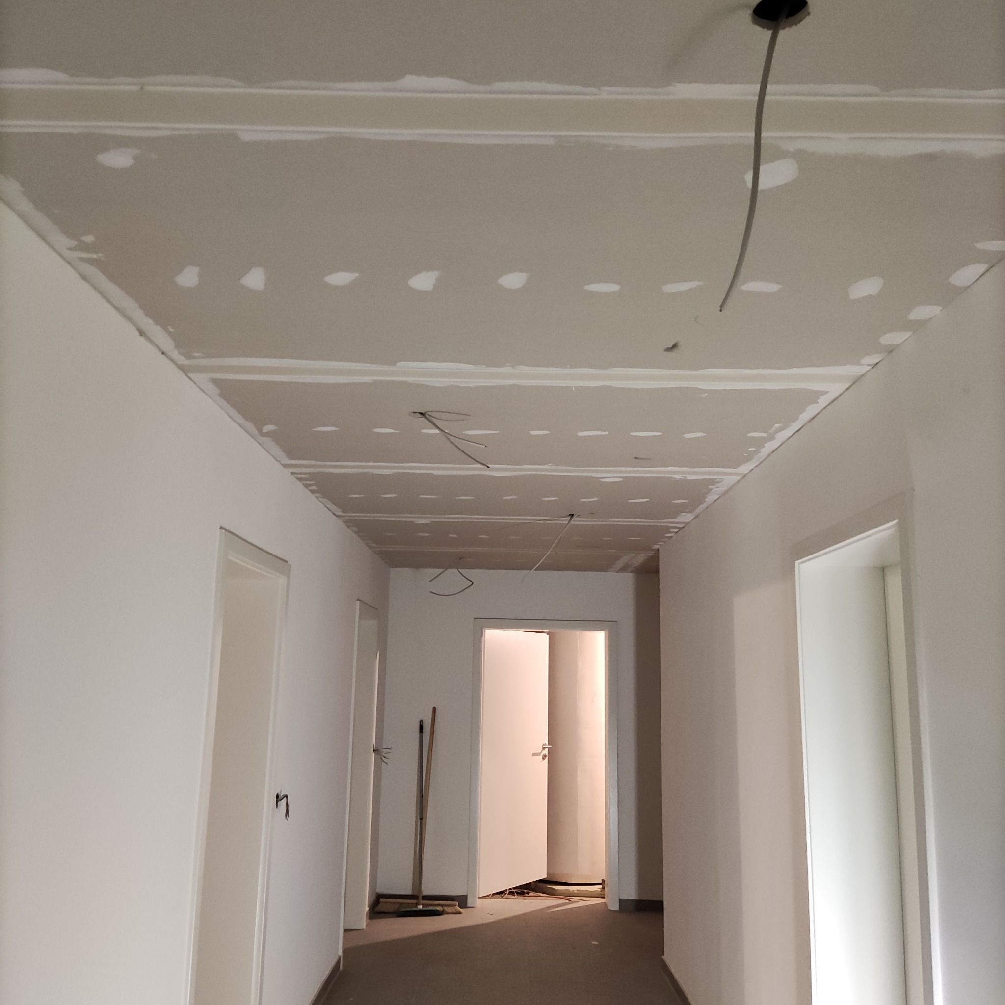Korytarz w trakcie remontu: sufit z płyt gipsowo-kartonowych z widocznymi łączeniami i przewodami elektrycznymi, ściany świeżo pomalowane na biało, otwarte drzwi w perspektywie.