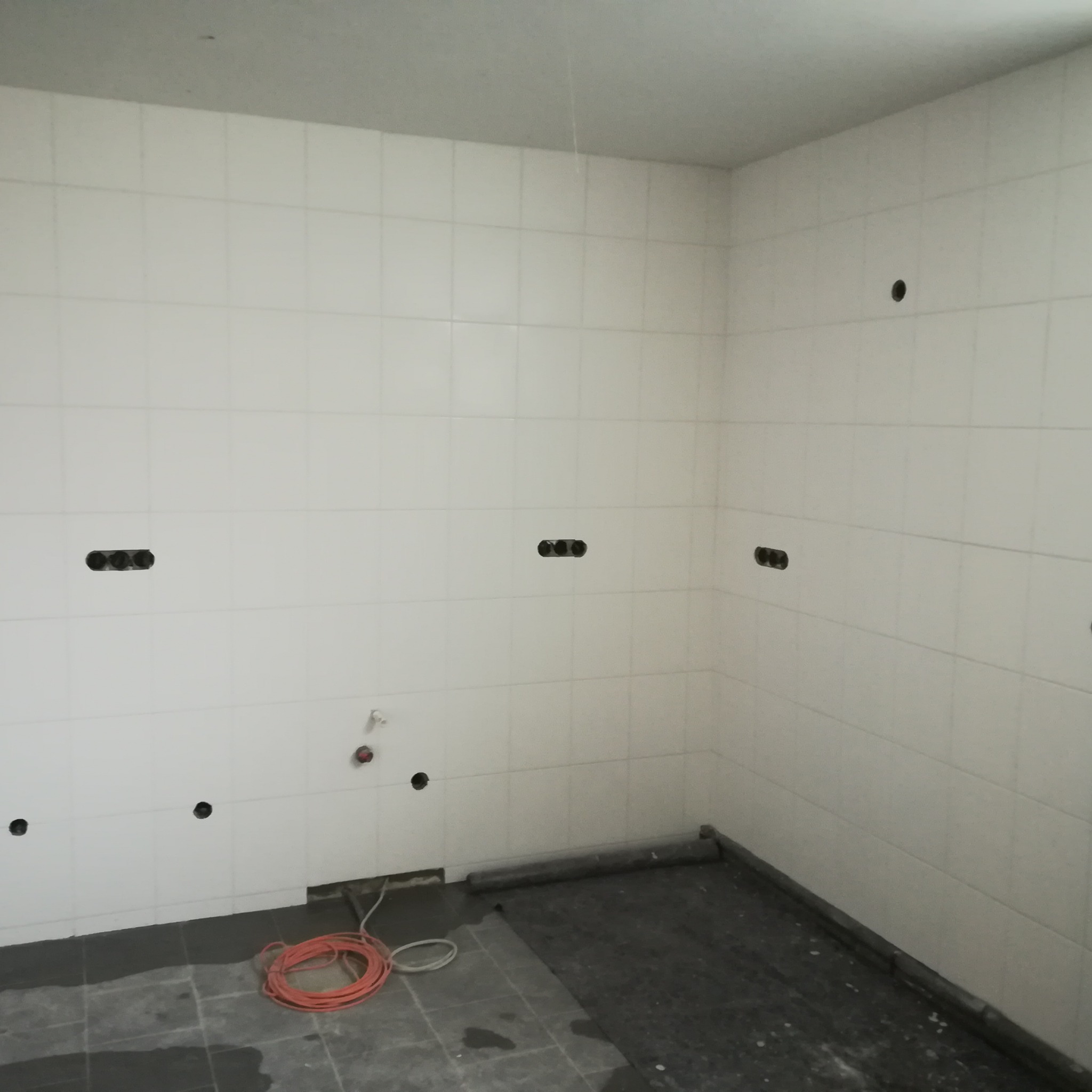 Ściana łazienki wyłożona białymi płytkami z przygotowanymi otworami na instalacje elektryczne i hydrauliczne, na podłodze folia ochronna i zwój pomarańczowego kabla.