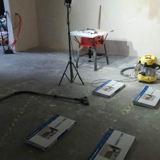 Przygotowania do montażu paneli: paczki paneli, odkurzacz przemysłowy, piła stołowa na statywie w surowym pomieszczeniu.