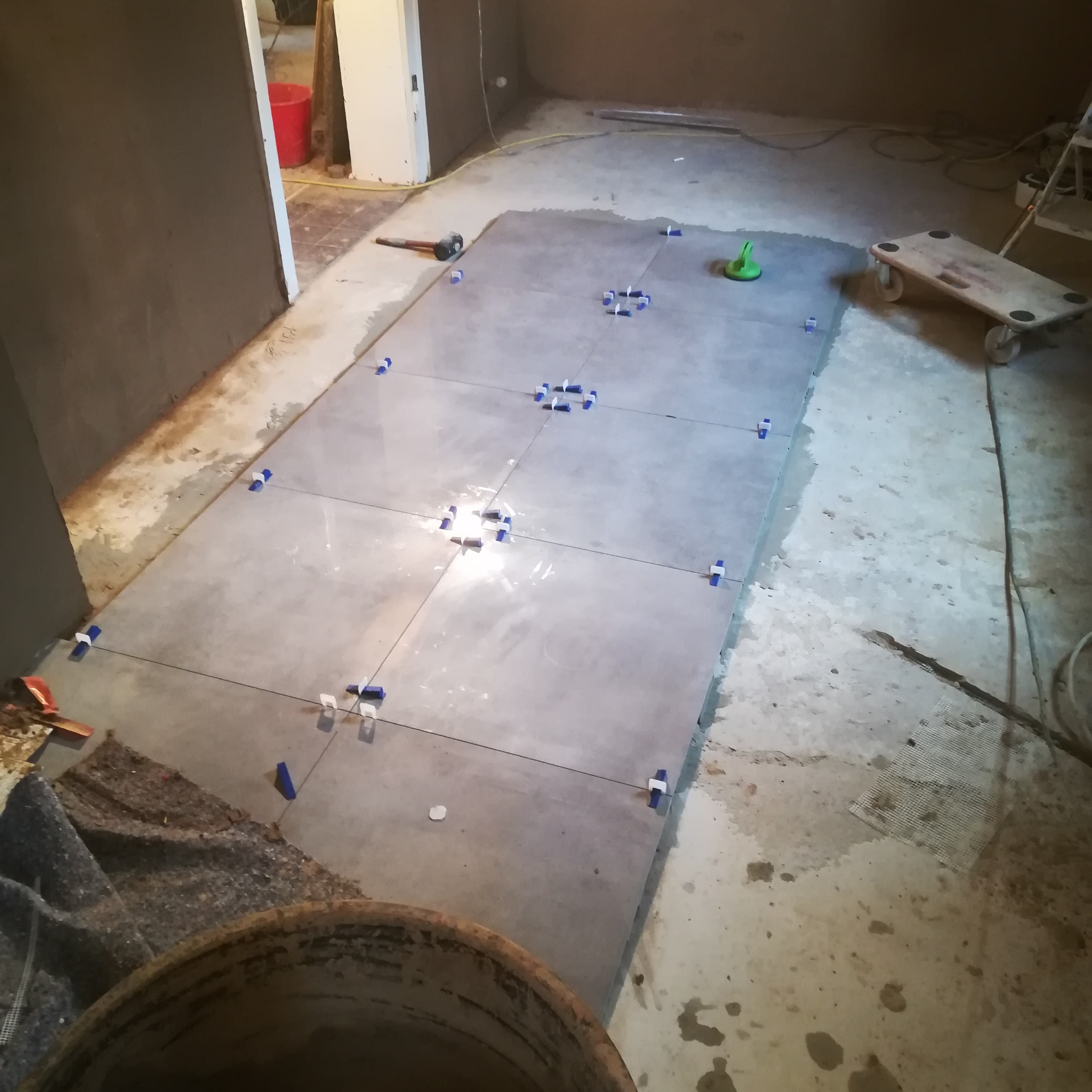 Układanie szarych płytek podłogowych w pomieszczeniu, widoczne krzyżyki dystansowe i narzędzia glazurnicze, w tle surowa ściana i wiadro z zaprawą.