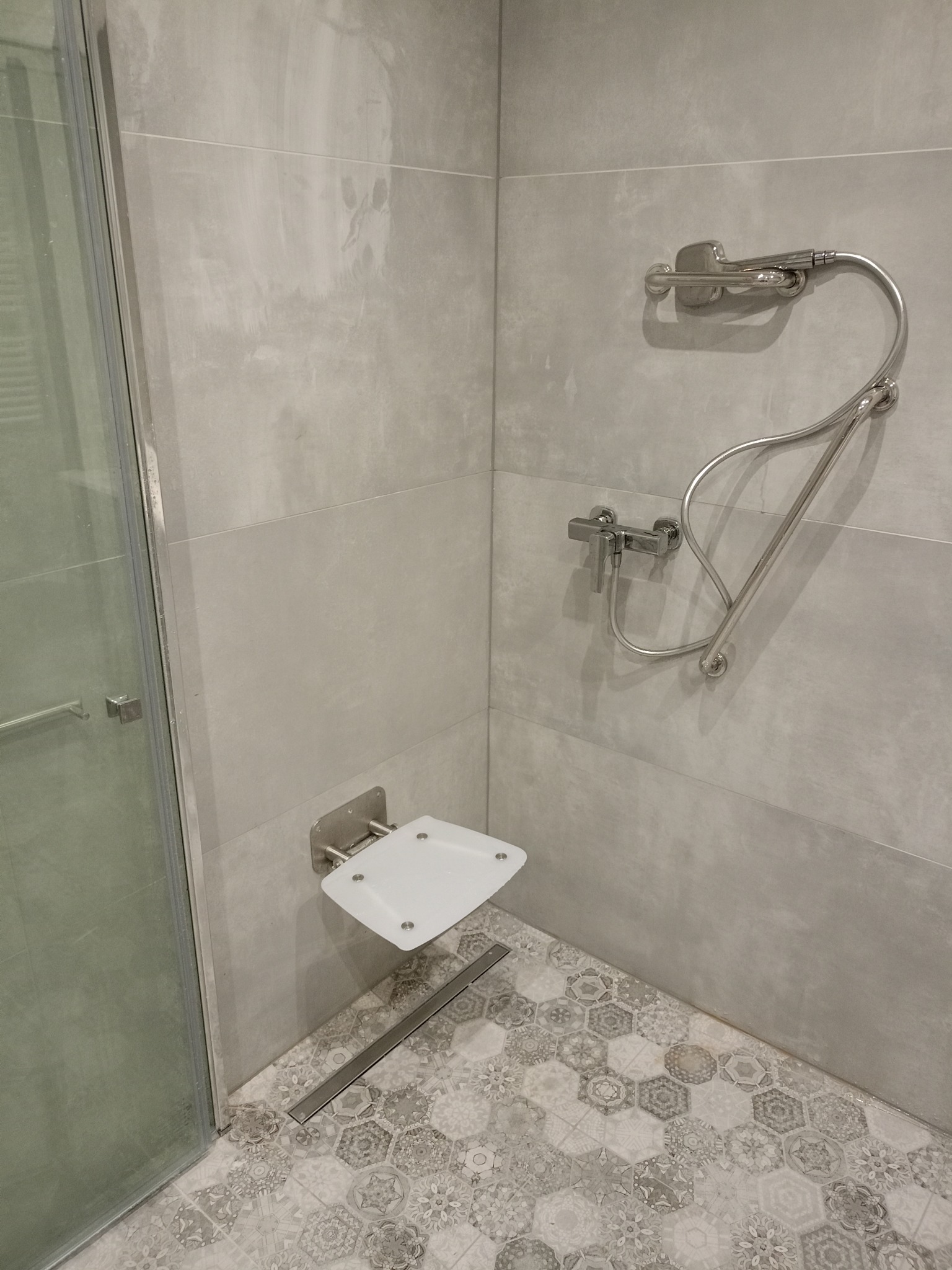 Nowoczesna kabina prysznicowa z siedziskiem i odpływem liniowym. Szare płytki ścienne i podłogowe w geometryczne wzory. Chromowana armatura i szklane drzwi.