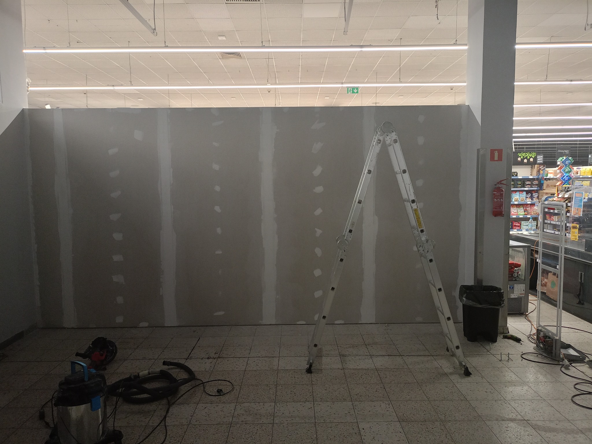 Montaż ścianki działowej z płyt karton-gips w przestrzeni handlowej. Widoczna drabina, szpachlowane łączenia płyt, narzędzia i odkurzacz przemysłowy. Oświetlenie sufitowe.