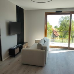 PROFIHOME & OLEKSANDR MELESHKO - Jasne wnętrze salonu z sofą, telewizorem na ścianie z dekoracyjnym panelem, widokiem na balkon i przyległą sypialnię.
