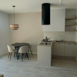 PROFIHOME & OLEKSANDR MELESHKO - Jasne wnętrze mieszkania z otwartą kuchnią, okrągły stół z krzesłami, widok na balkon i zabudowę kuchenną z białymi szafkami i czarnym okapem.