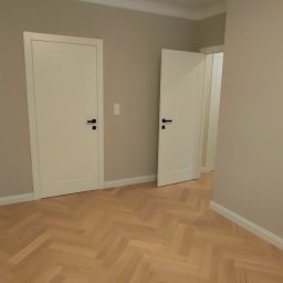 PROFIHOME & OLEKSANDR MELESHKO - Pusty pokój z parkietem ułożonym w jodełkę, dwoje białych drzwi z czarnymi klamkami, kremowe ściany i białe listwy przypodłogowe.