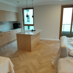 PROFIHOME & OLEKSANDR MELESHKO - Jasna kuchnia z wyspą i parkietem w jodełkę, częściowo zakryte meble folią ochronną.