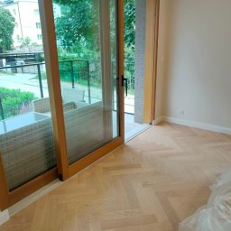 PROFIHOME & OLEKSANDR MELESHKO - Jasny parkiet ułożony w jodełkę francuską, przesuwne drzwi balkonowe z drewnianą ramą otwarte na taras z meblami ogrodowymi.