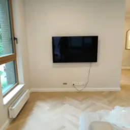 Salon po remoncie z parkietem ułożonym w jodełkę, zamontowanym telewizorem i grzejnikiem pod oknem.