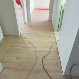 PROFIHOME & OLEKSANDR MELESHKO - Wnętrze w trakcie prac wykończeniowych: jasne panele podłogowe, białe ściany, widoczne kable elektryczne i fragment sylwetki pracownika w oddali.