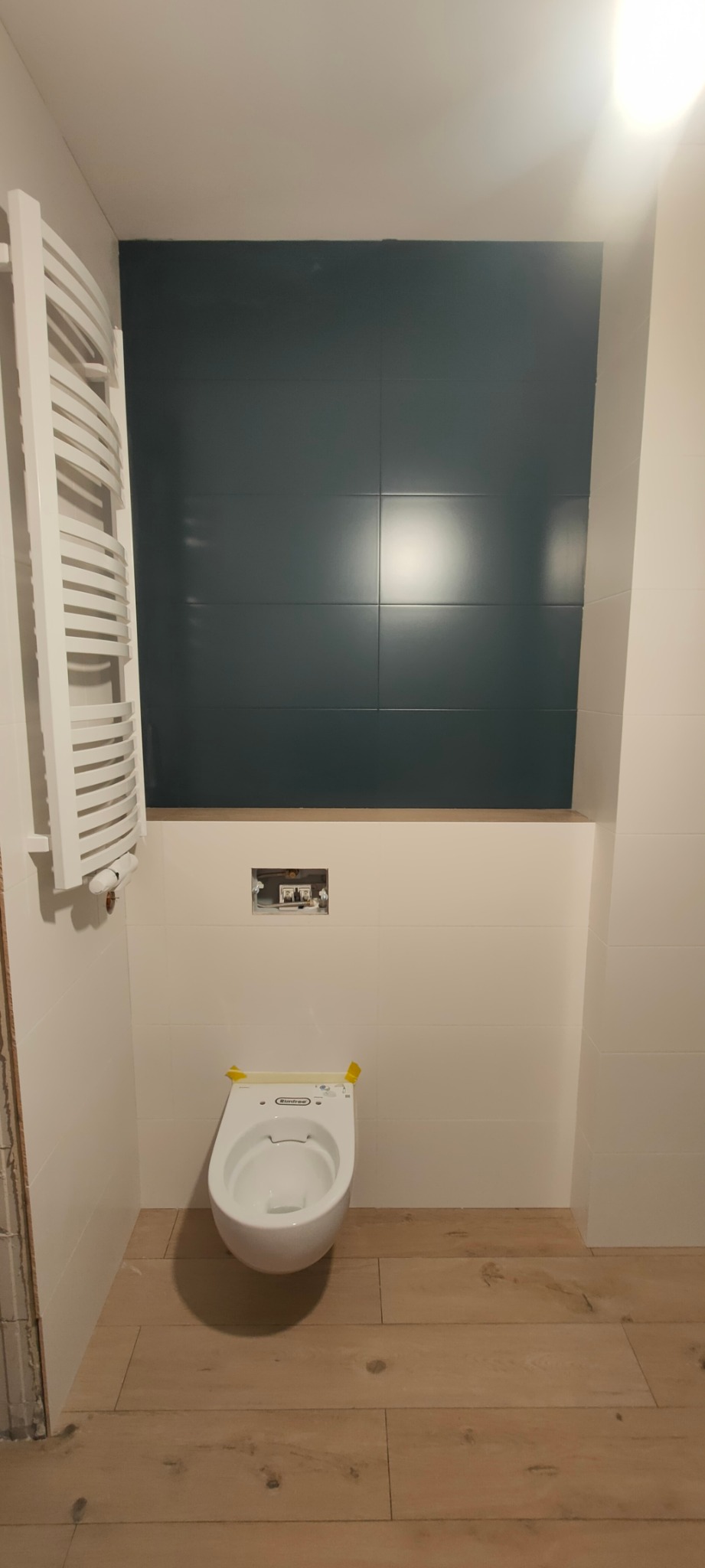 Wisząca toaleta z żółtą taśmą zabezpieczającą, zamontowana na podłodze z paneli imitujących drewno, z fragmentem ściany wyłożonej ciemnozielonymi płytkami i białym grzejnikiem drabinkowym po lewej...