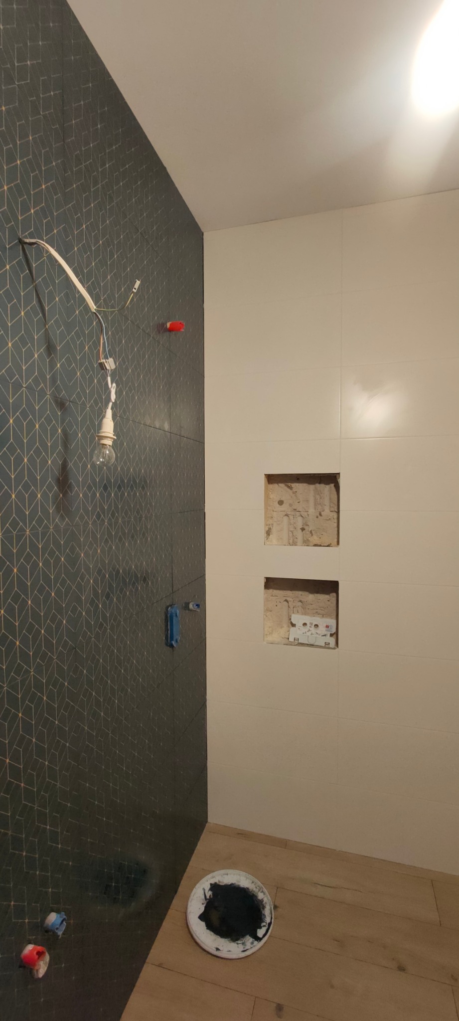 Łazienka w trakcie wykańczania w Bukowcu: ułożona część glazury z geometrycznym wzorem, białe płytki z wnękami, wiszące kable z żarówką i wiadro z farbą na podłodze.