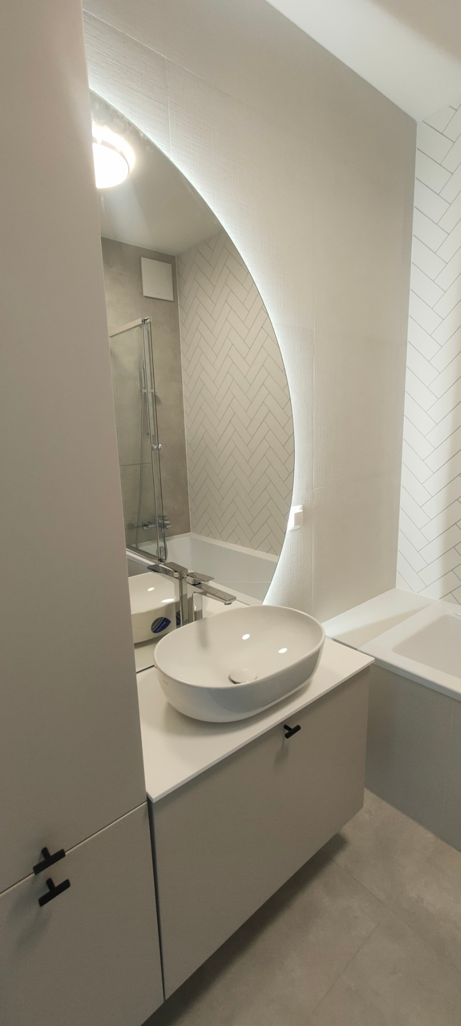 Nowoczesna łazienka z owalną umywalką nablatową, podświetlanym lustrem i białymi płytkami ułożonymi w jodełkę.