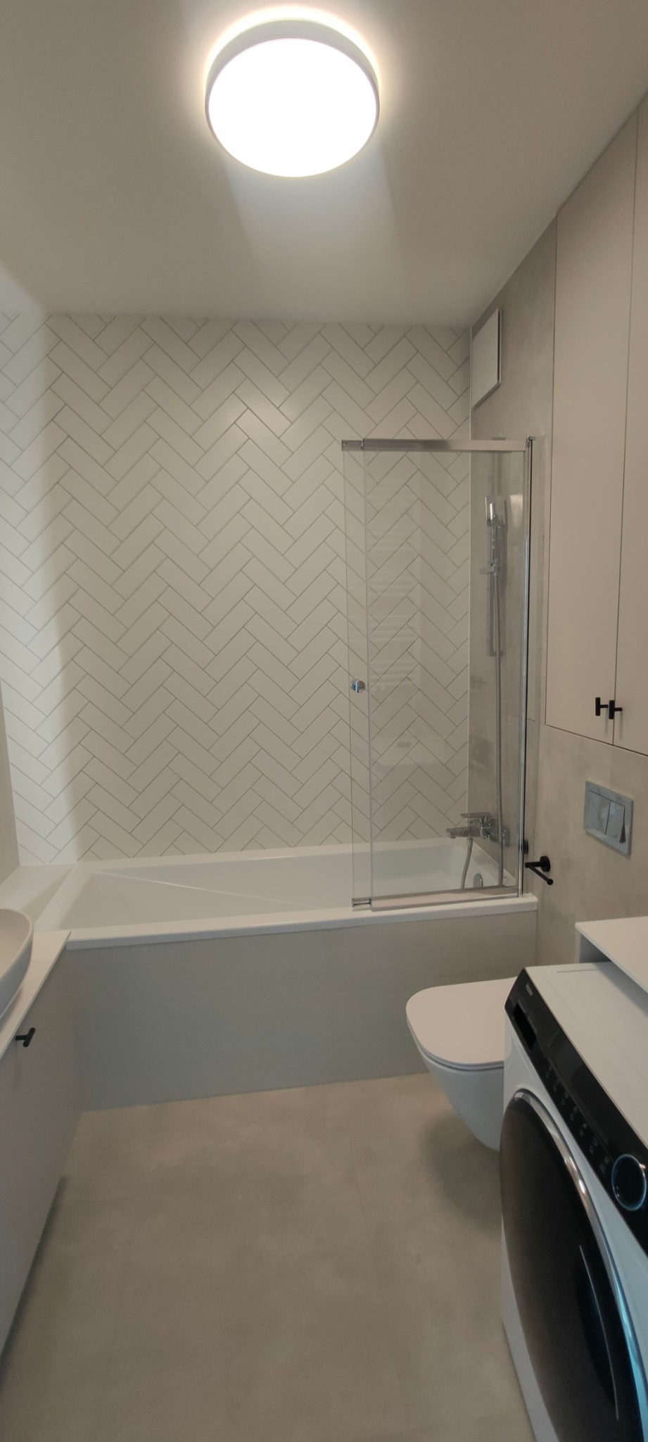 Nowoczesna łazienka po remoncie z białą armaturą, płytkami ułożonymi w jodełkę na ścianie, kabiną nawannową i pralką.