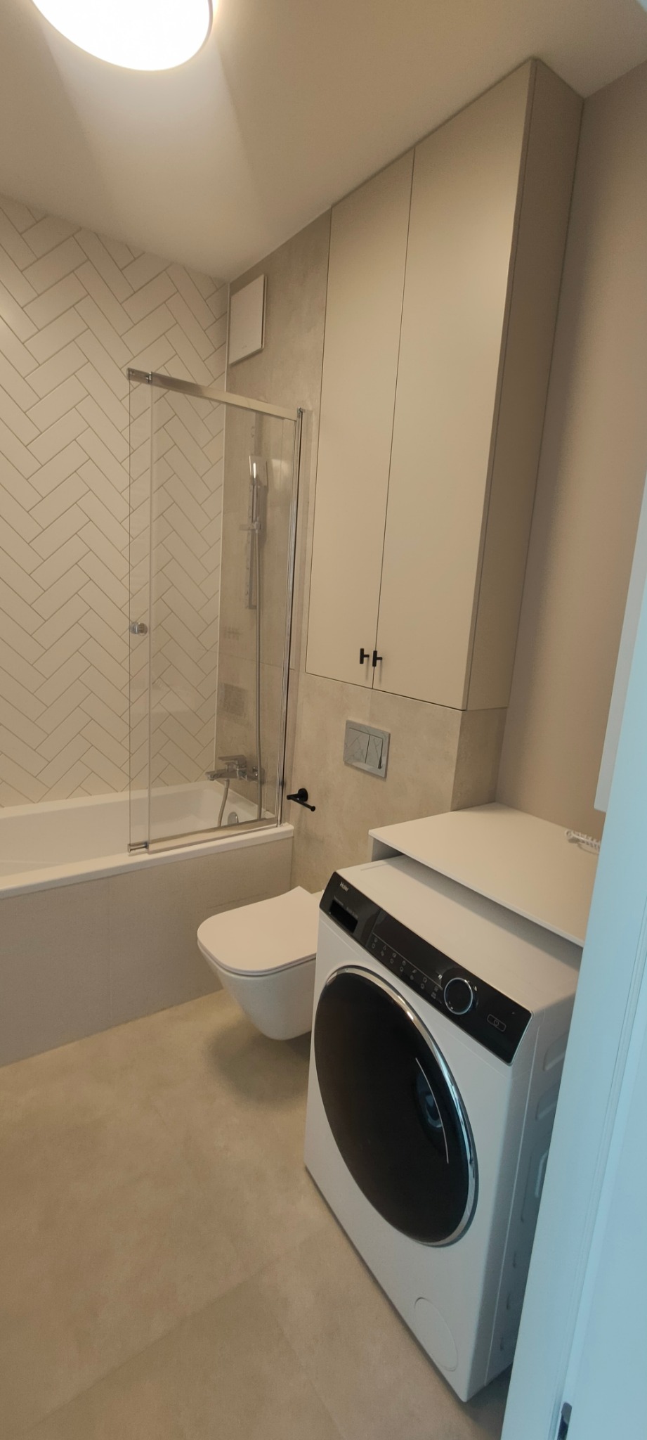 Nowoczesna łazienka z białą armaturą, płytkami w jodełkę i pralką z czarnym panelem, zabudowana szafka nad toaletą.