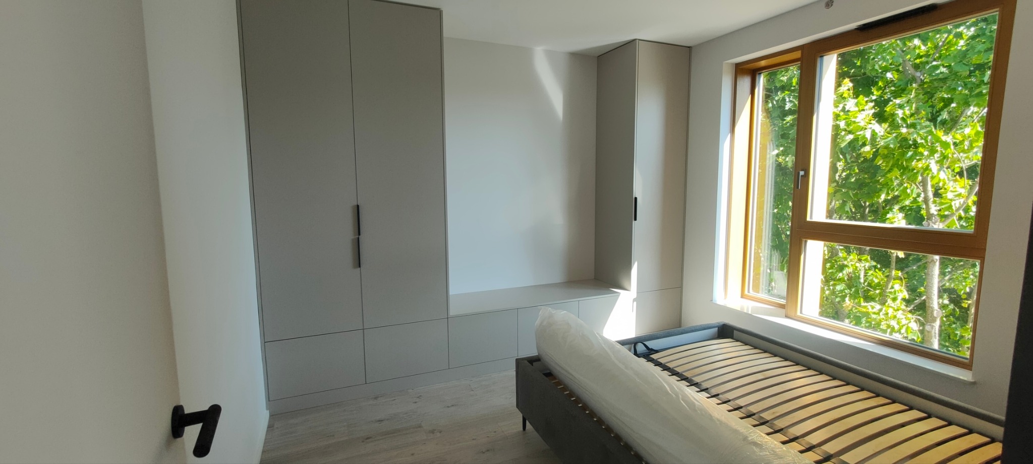 Wnętrze pokoju z zabudową meblową w odcieniach szarości, łóżko z drewnianym stelażem i widokiem na zieleń przez okno.