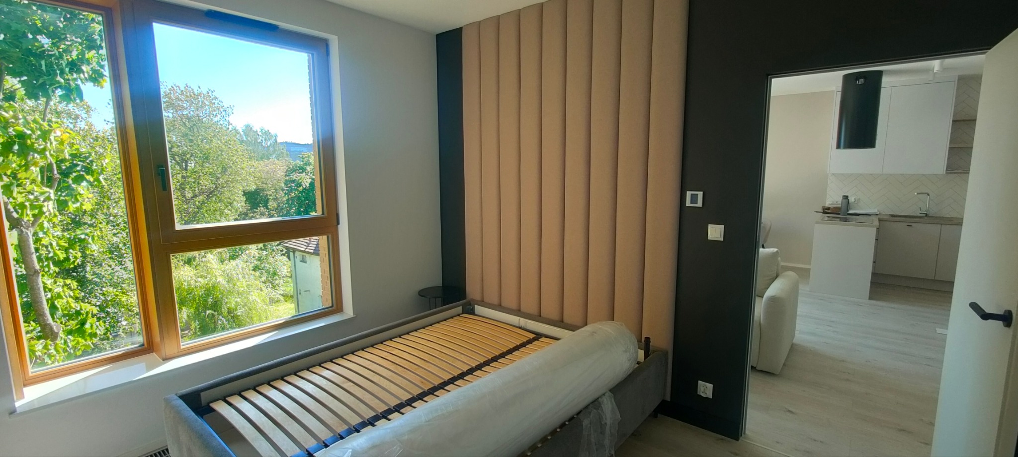 Pusta rama łóżka z drewnianym stelażem i beżowym panelem ściennym w nowym pokoju, widok na zielone drzewa za oknem i fragment kuchni w tle.