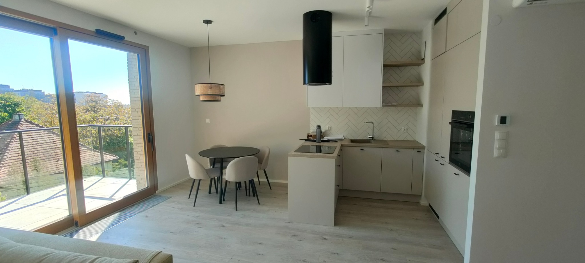 Jasne wnętrze mieszkania z otwartą kuchnią, okrągły stół z krzesłami, widok na balkon i zabudowę kuchenną z białymi szafkami i czarnym okapem.