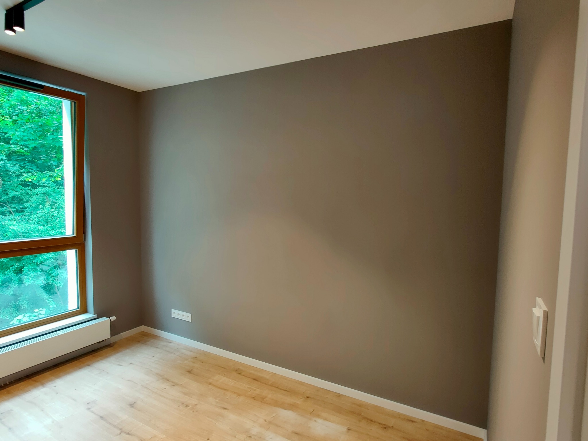 Pomalowane na szaro wnętrze pokoju z drewnianą podłogą, widok na zieleń za oknem, biały grzejnik i gniazdko elektryczne na ścianie.