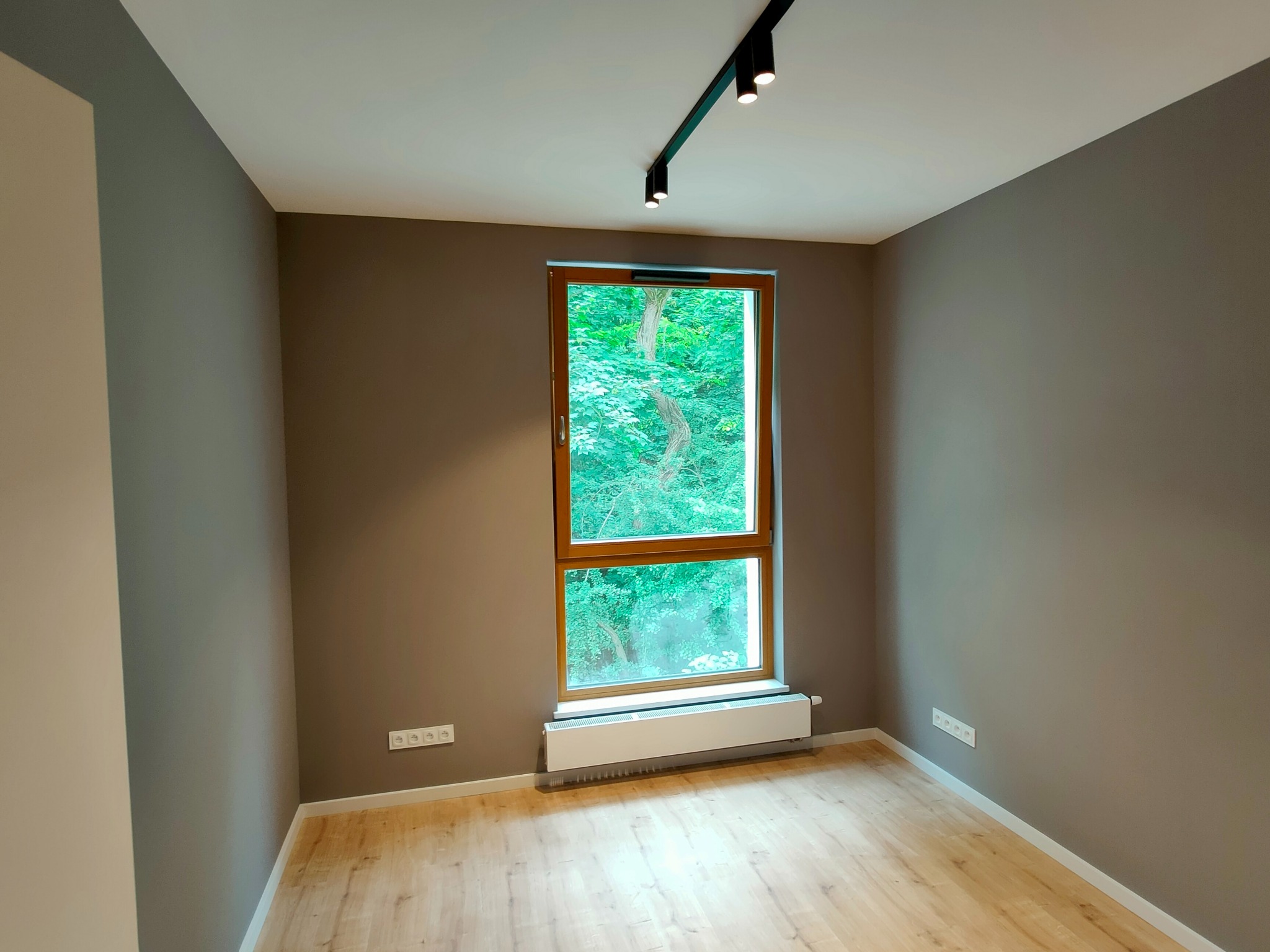 Pusty pokój z drewnianą podłogą, szarobrązowymi ścianami, oknem z widokiem na zieleń i nowoczesnym oświetleniem szynowym na suficie.