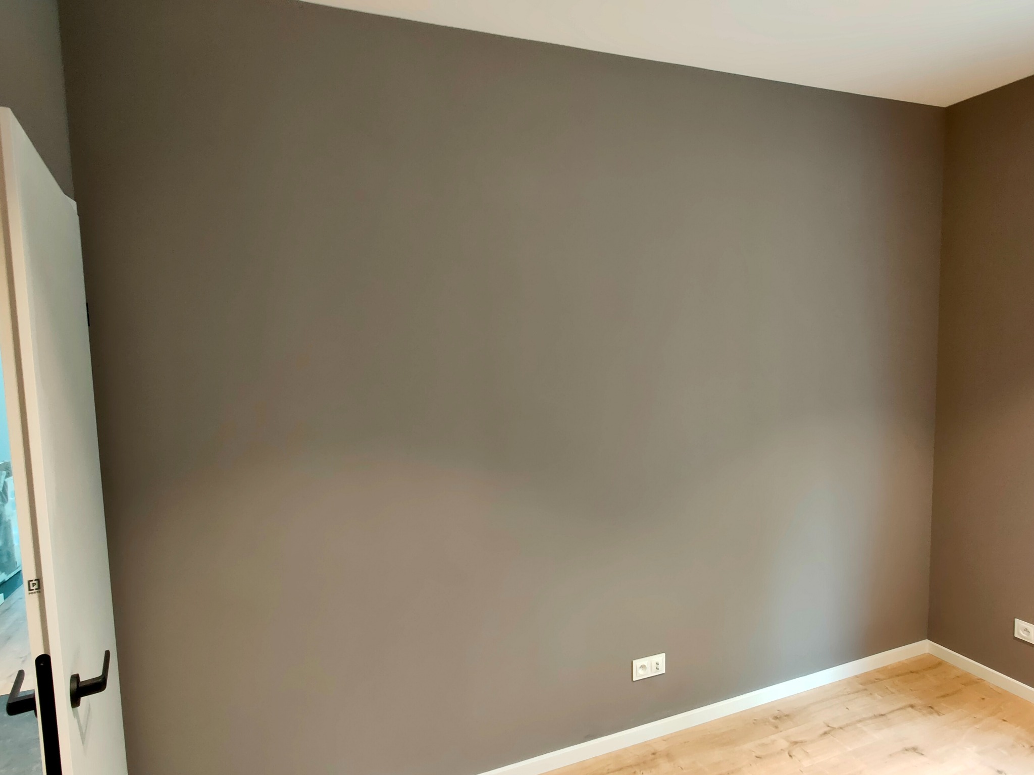 Pomalowane na szaro ściany w rogu pokoju z drewnianą podłogą i białymi listwami przypodłogowymi, widoczne gniazdka elektryczne i fragment białych drzwi z czarną klamką.