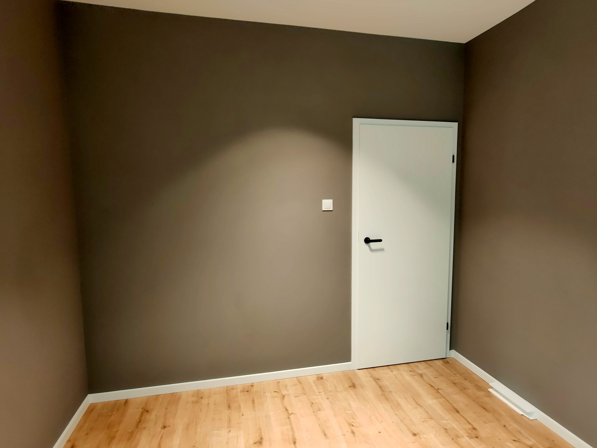 Pomalowane na brązowo ściany, białe listwy przypodłogowe i podłoga z paneli w pustym pokoju, białe drzwi z czarną klamką.