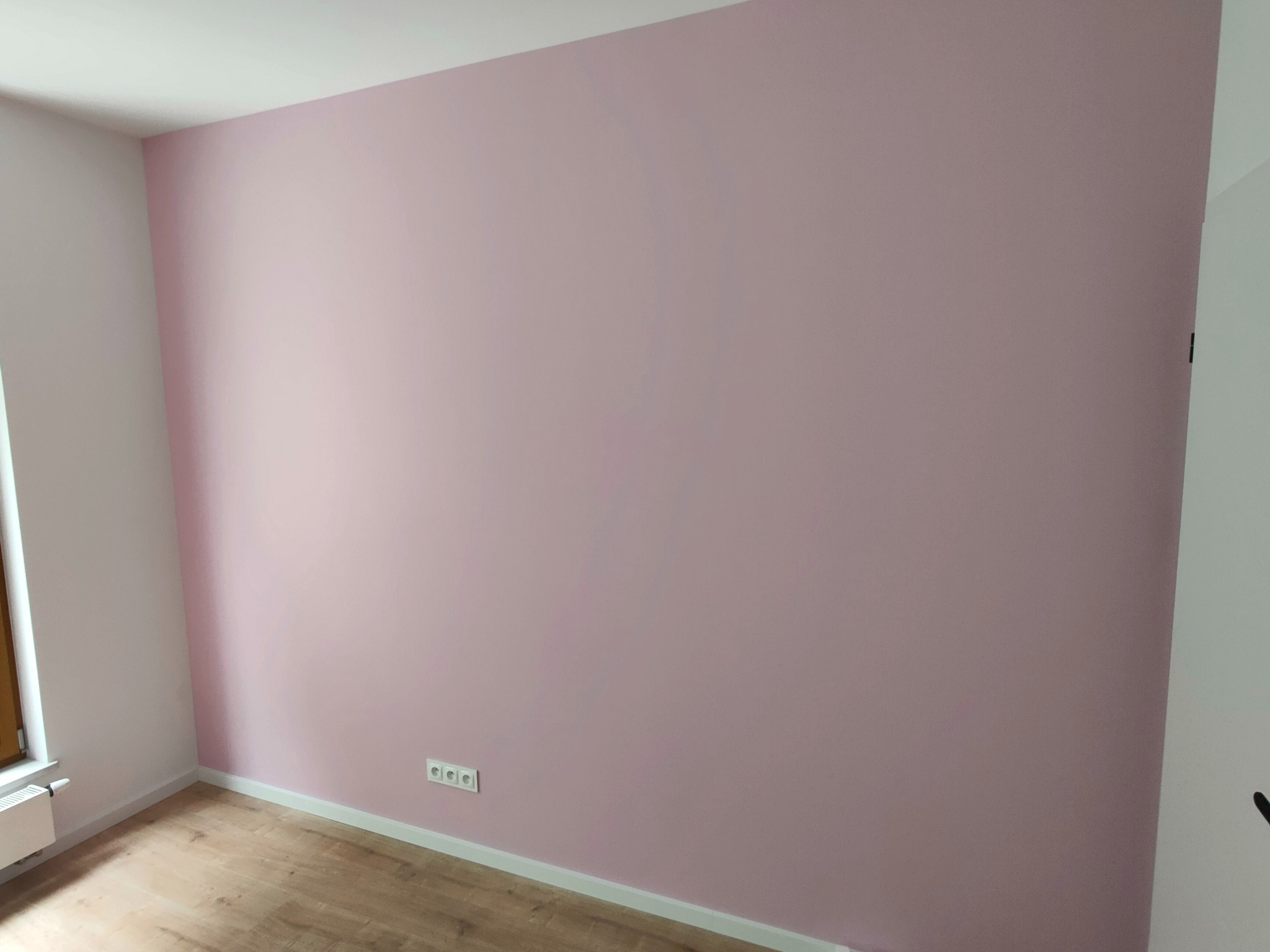 Pomalowana na różowo ściana w pokoju z drewnianą podłogą i białymi listwami przypodłogowymi, widoczne gniazdka elektryczne.