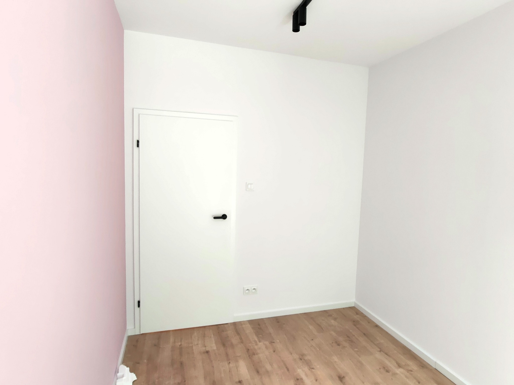 Wnętrze pokoju po remoncie: ściana pomalowana na różowo, białe ściany, drewnopodobna podłoga, białe drzwi z czarną klamką, czarna lampa sufitowa.