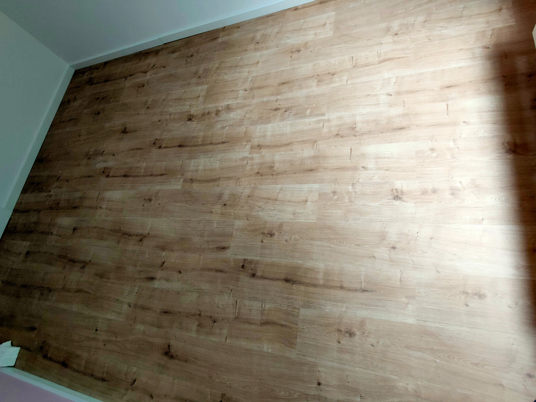 Ściana wykończona panelami imitującymi jasne drewno, widoczne łączenia paneli i listwa przypodłogowa.