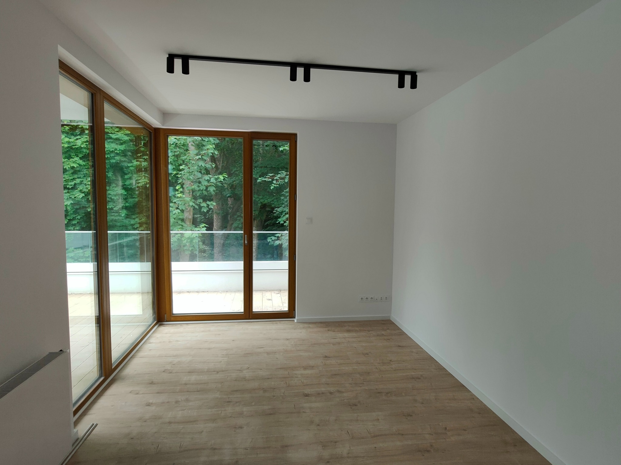 Pusty, jasny pokój z drewnianą podłogą, dużymi oknami balkonowymi z widokiem na zieleń i nowoczesnym oświetleniem sufitowym.