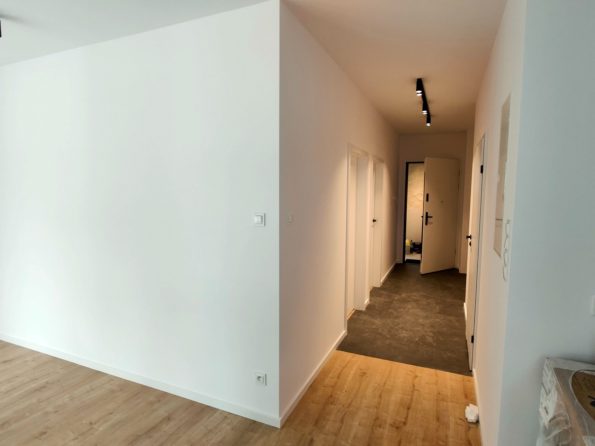 Wnętrze mieszkania po remoncie: białe ściany, jasna drewniana podłoga przechodząca w ciemny korytarz z trzema białymi drzwiami i czarnym oświetleniem szynowym.