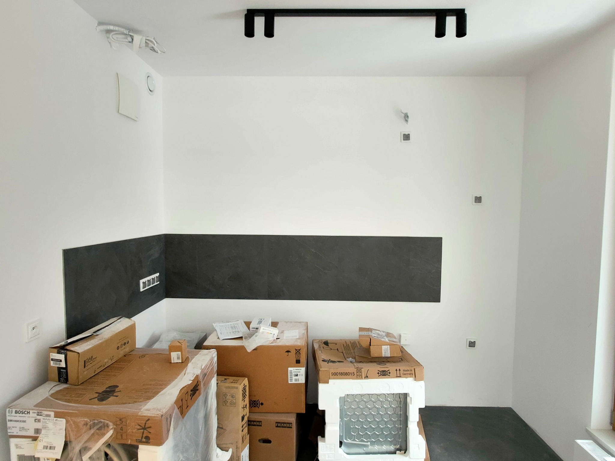 Wnętrze kuchni w trakcie prac wykończeniowych: widoczne kartony ze sprzętem AGD (Bosch, Franke) pod ścianą z fragmentem położonych ciemnych płytek, surowa instalacja elektryczna na ścianie i suficie.