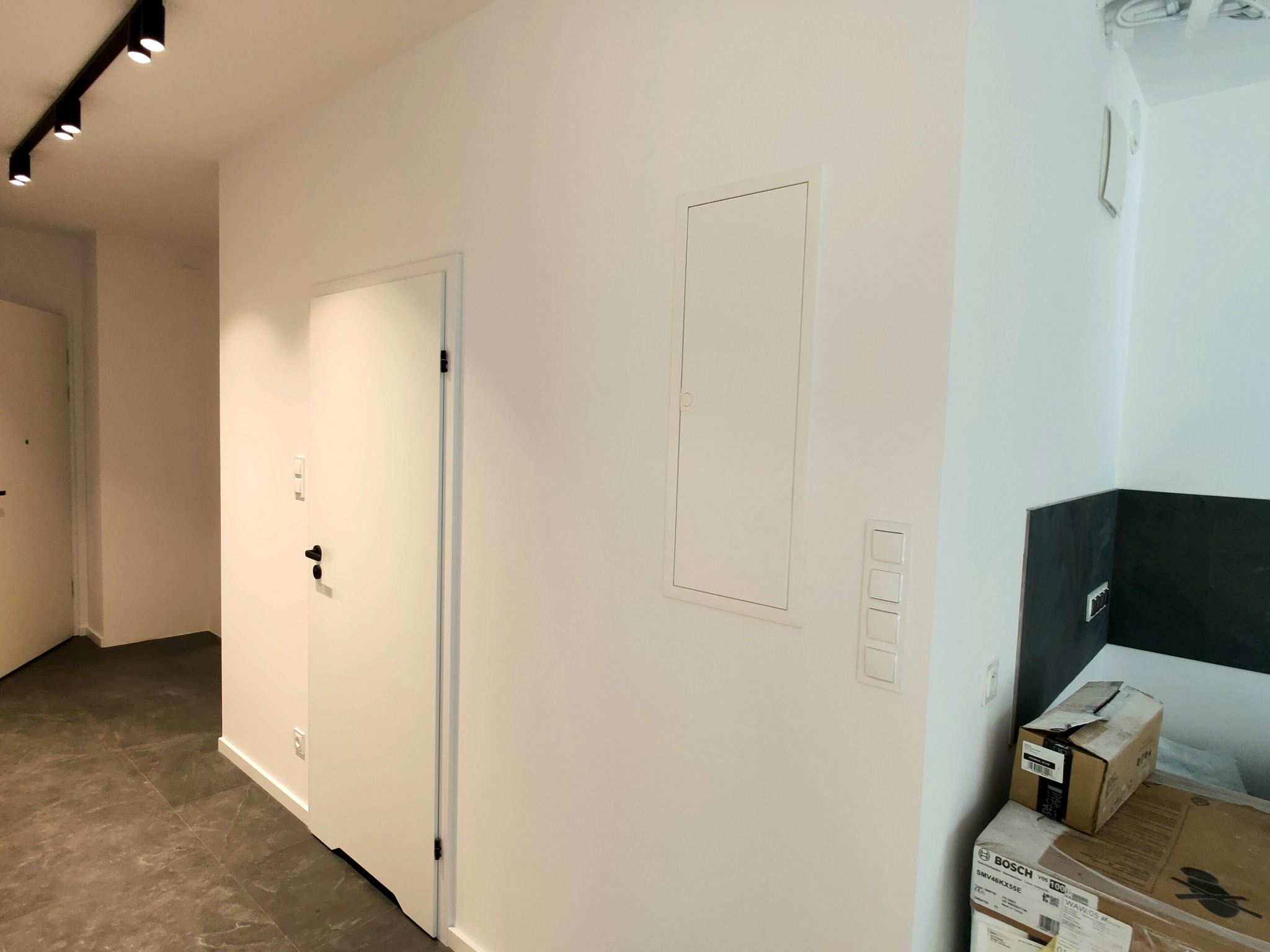 Wnętrze mieszkania po remoncie: białe ściany, szara podłoga z płytek, białe drzwi z czarną klamką, skrzynka rewizyjna na ścianie, czarna listwa oświetleniowa na suficie, kartony z urządzeniami AGD...