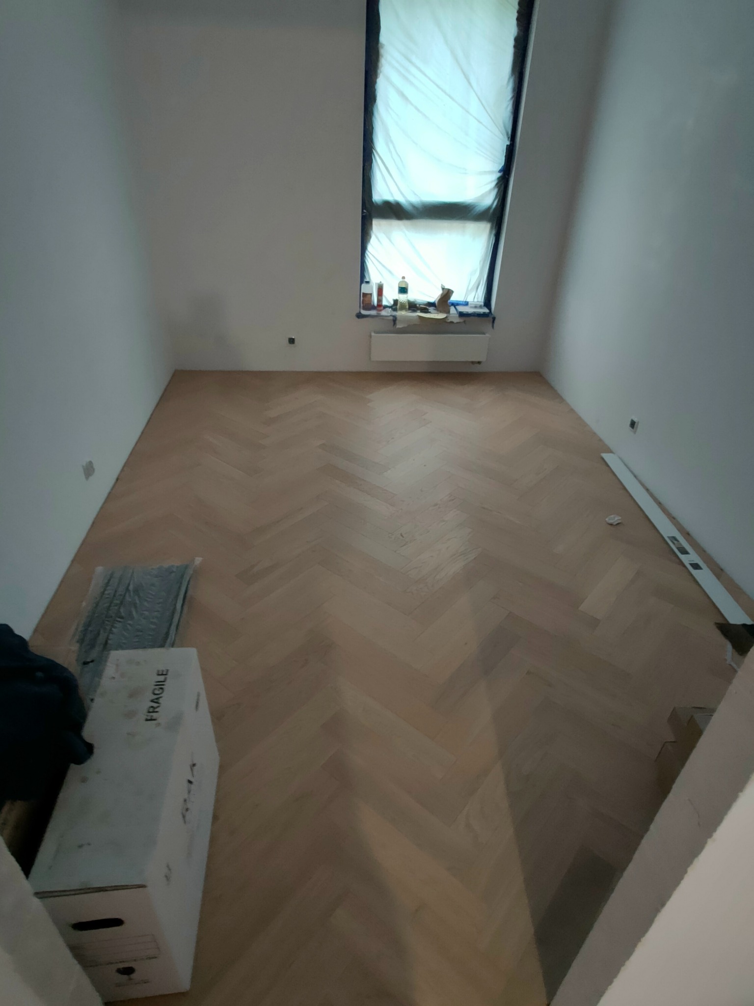 Ułożona parkietowa podłoga w jodełkę w pustym, jasnym pokoju, okno zabezpieczone folią, widoczny karton z napisem 'FRAGILE' i poziomica.