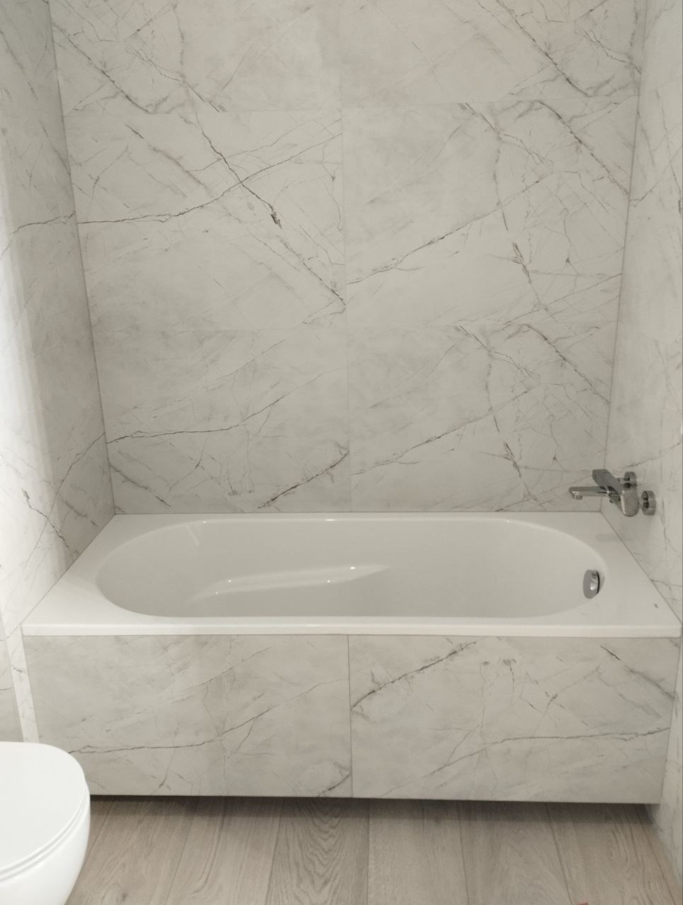 Biała wanna obudowana płytkami imitującymi marmur, z widoczną chromowaną baterią i fragmentem toalety, na tle ściany wyłożonej identycznymi płytkami.