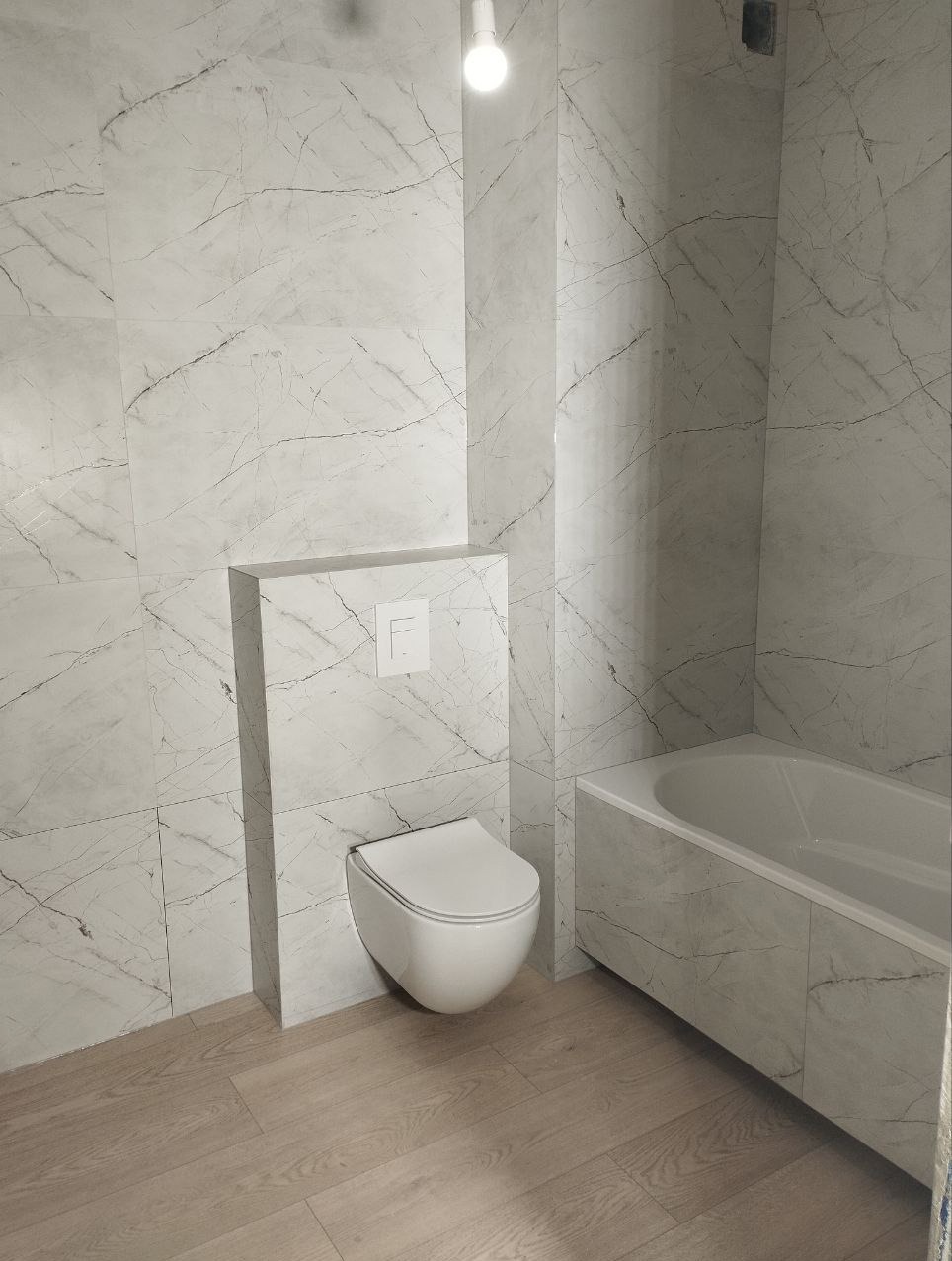 Nowoczesna łazienka wykończona płytkami imitującymi marmur, z podwieszaną toaletą i zabudowaną wanną, jasna podłoga z paneli.
