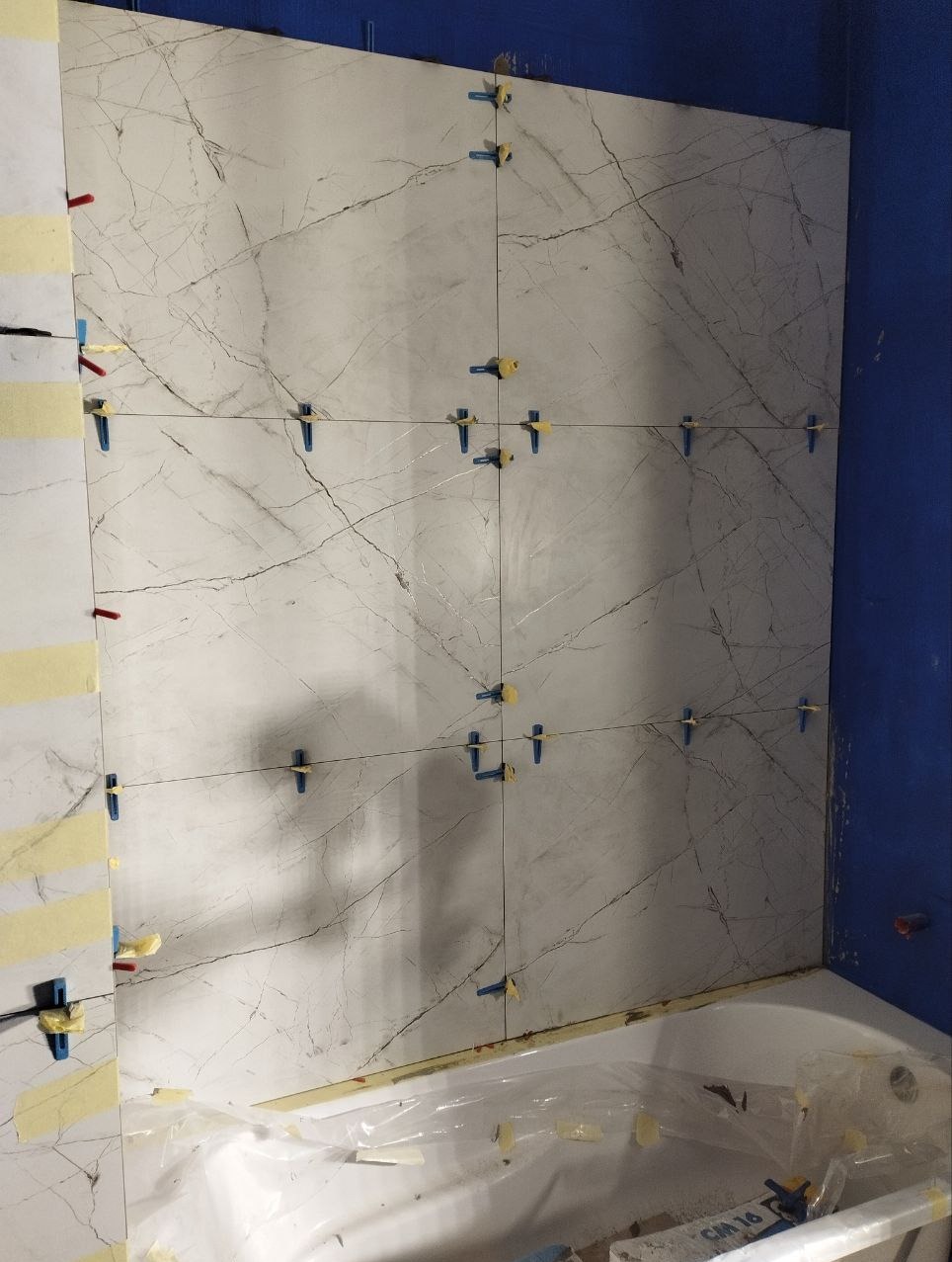 Układanie marmurowych płytek w łazience, widoczne krzyżyki dystansowe i taśma malarska zabezpieczająca wannę.