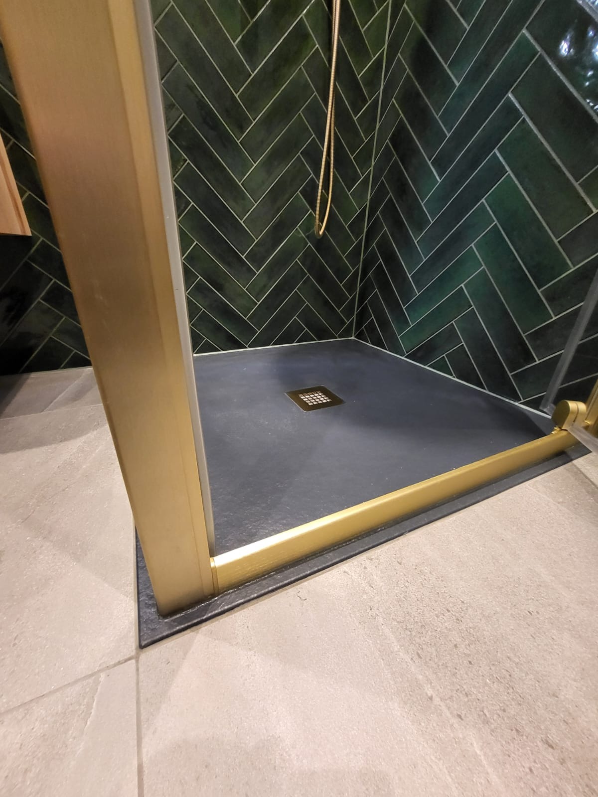 Kabina prysznicowa z ciemnozielonymi płytkami ułożonymi w jodełkę i złotymi detalami. Widoczny odpływ liniowy i fragment podłogi wyłożonej jasnymi płytkami.