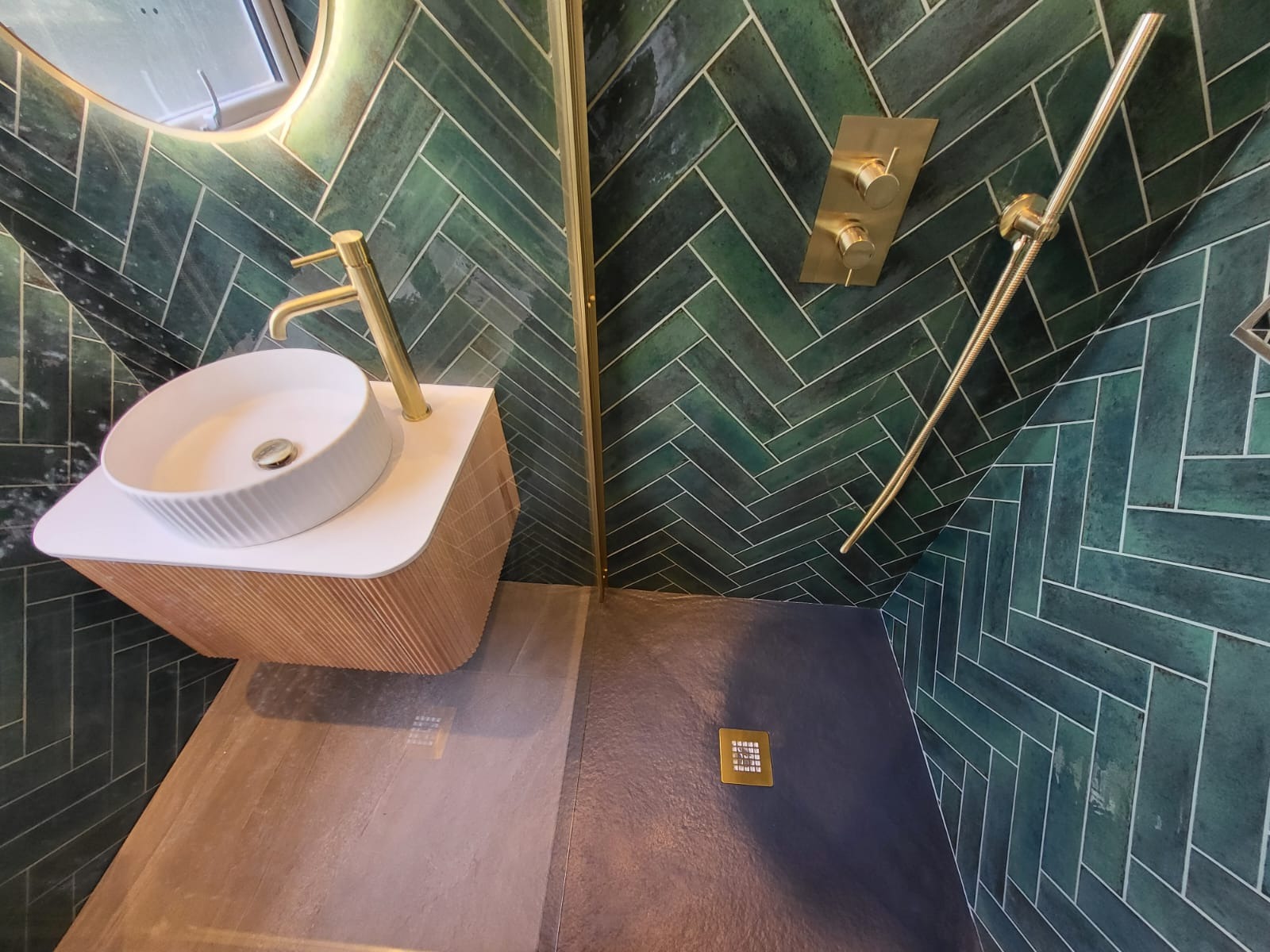Nowoczesna łazienka z zielonymi płytkami ułożonymi w jodełkę, złotą armaturą i umywalką na drewnianej szafce. Widoczny odpływ liniowy i oświetlenie lustra.