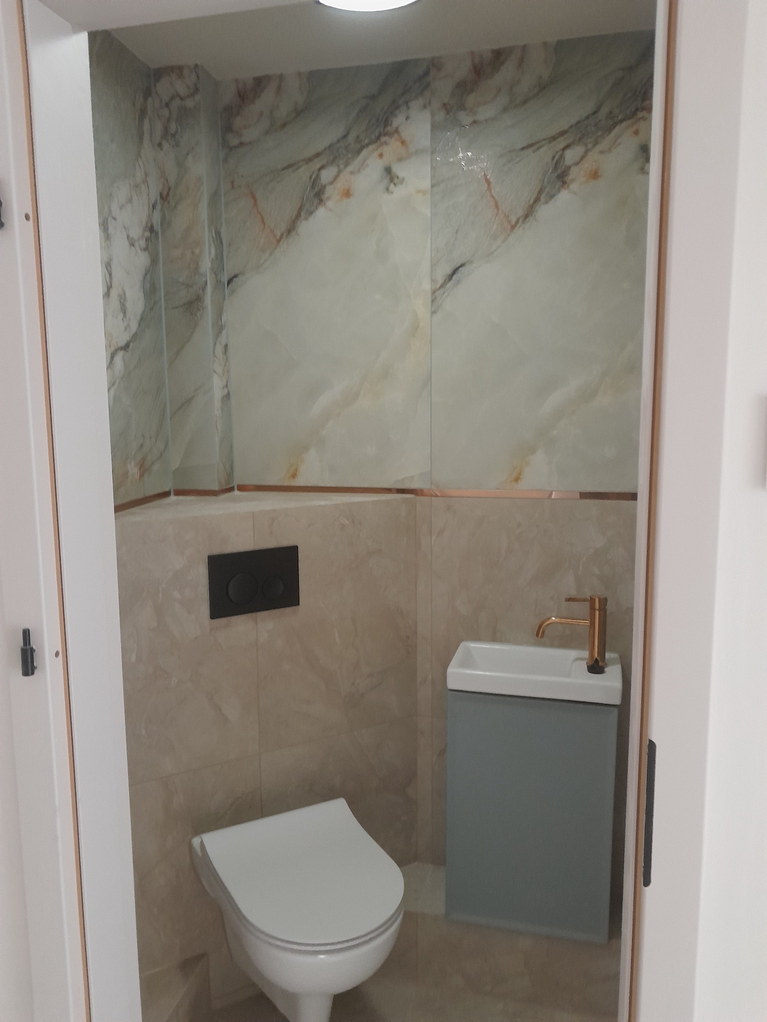 Kompaktowa łazienka z marmurowymi płytkami, podwieszaną toaletą i umywalką z szafką w odcieniach szarości. Złota bateria dodaje elegancji. Widok zza futryny.