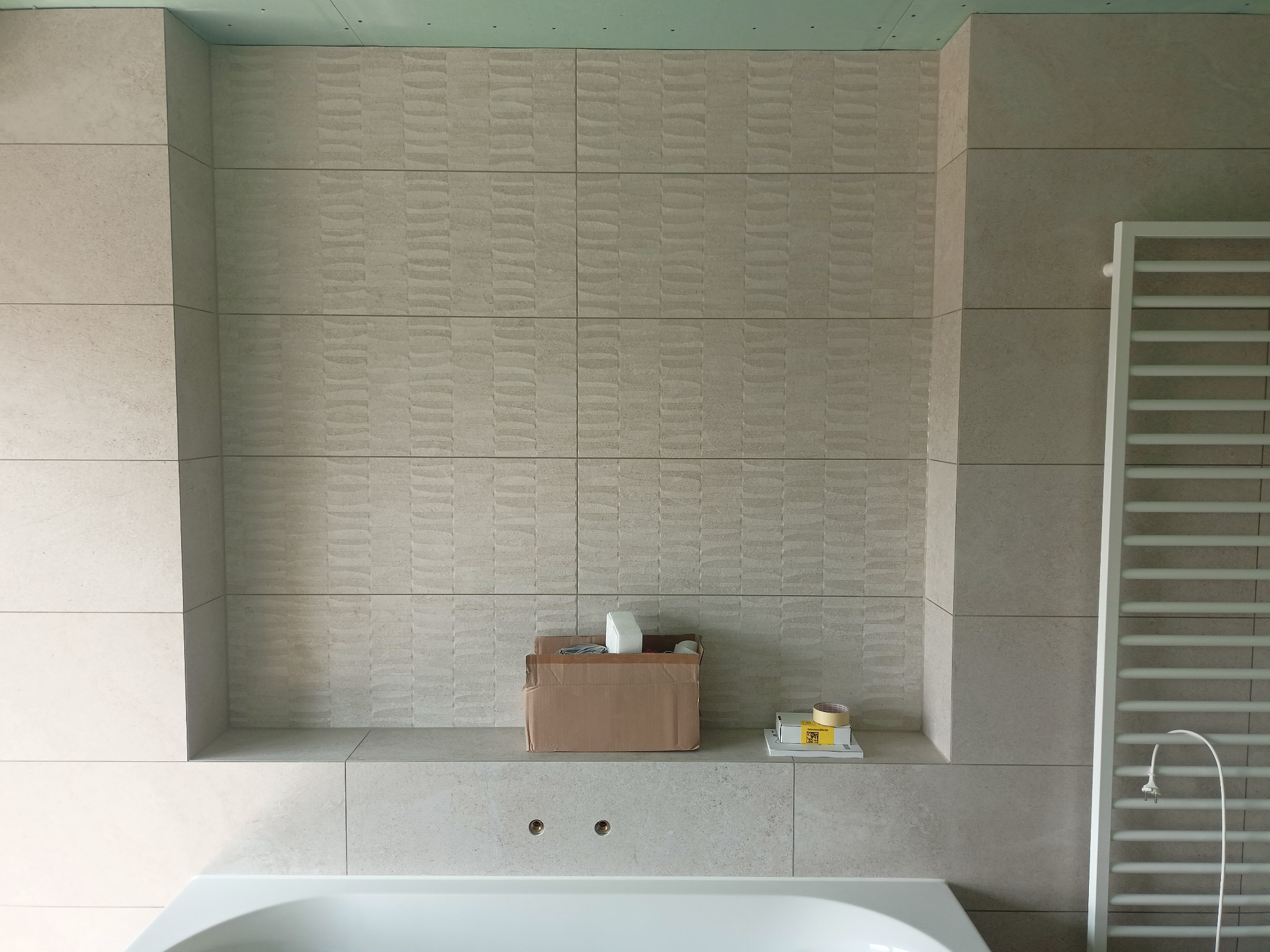 Ściana w łazience wyłożona beżowymi płytkami z delikatnym wzorem, widoczna wanna, grzejnik drabinkowy z wystającym kablem i karton z akcesoriami na półce, sufit w kolorze miętowym.
