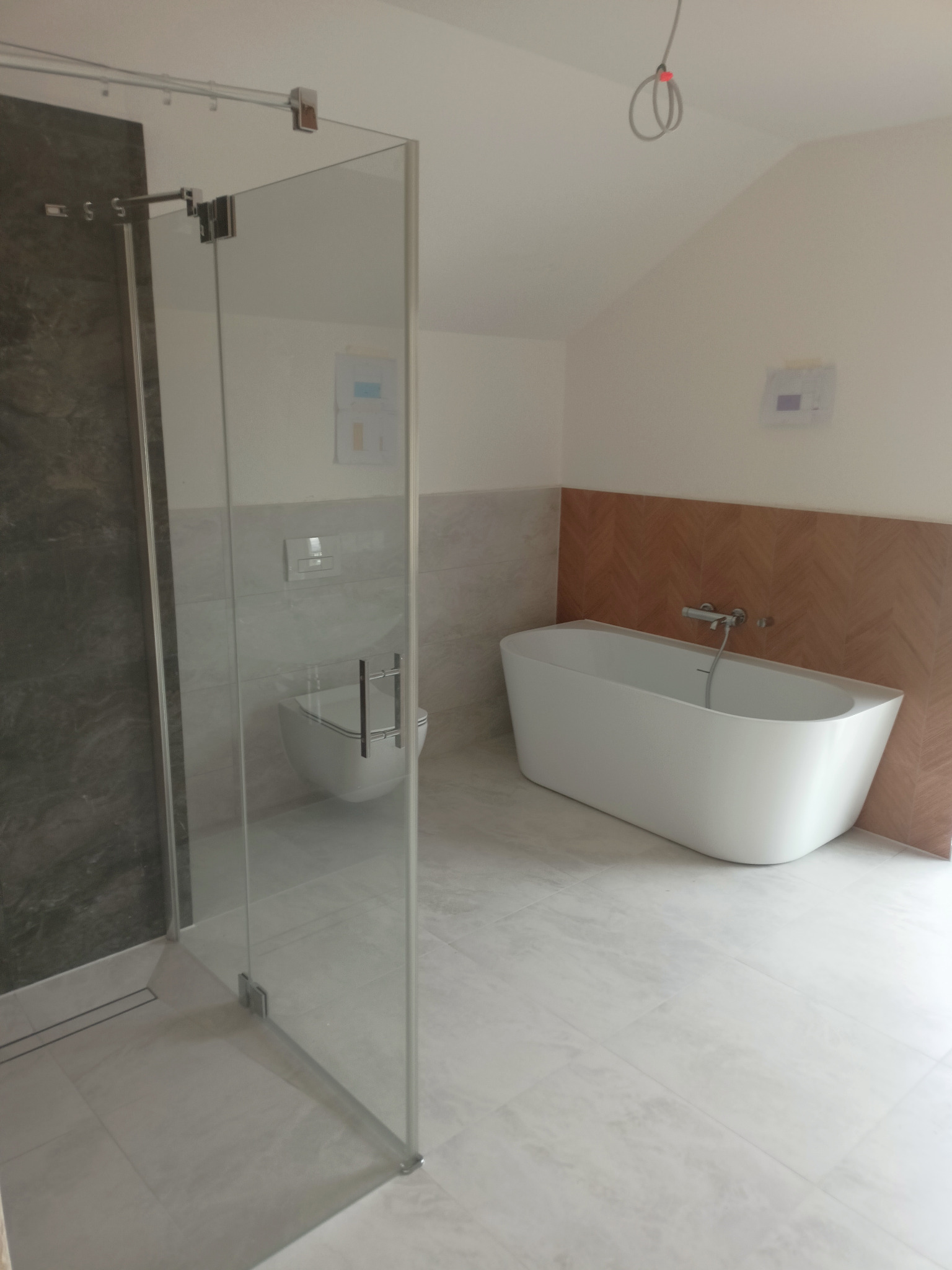 Nowoczesna łazienka w Białymstoku z kabiną prysznicową typu walk-in z szarymi płytkami imitującymi kamień, białą wiszącą toaletą i wolnostojącą wanną na tle ściany wykończonej drewnopodobną mozaiką.