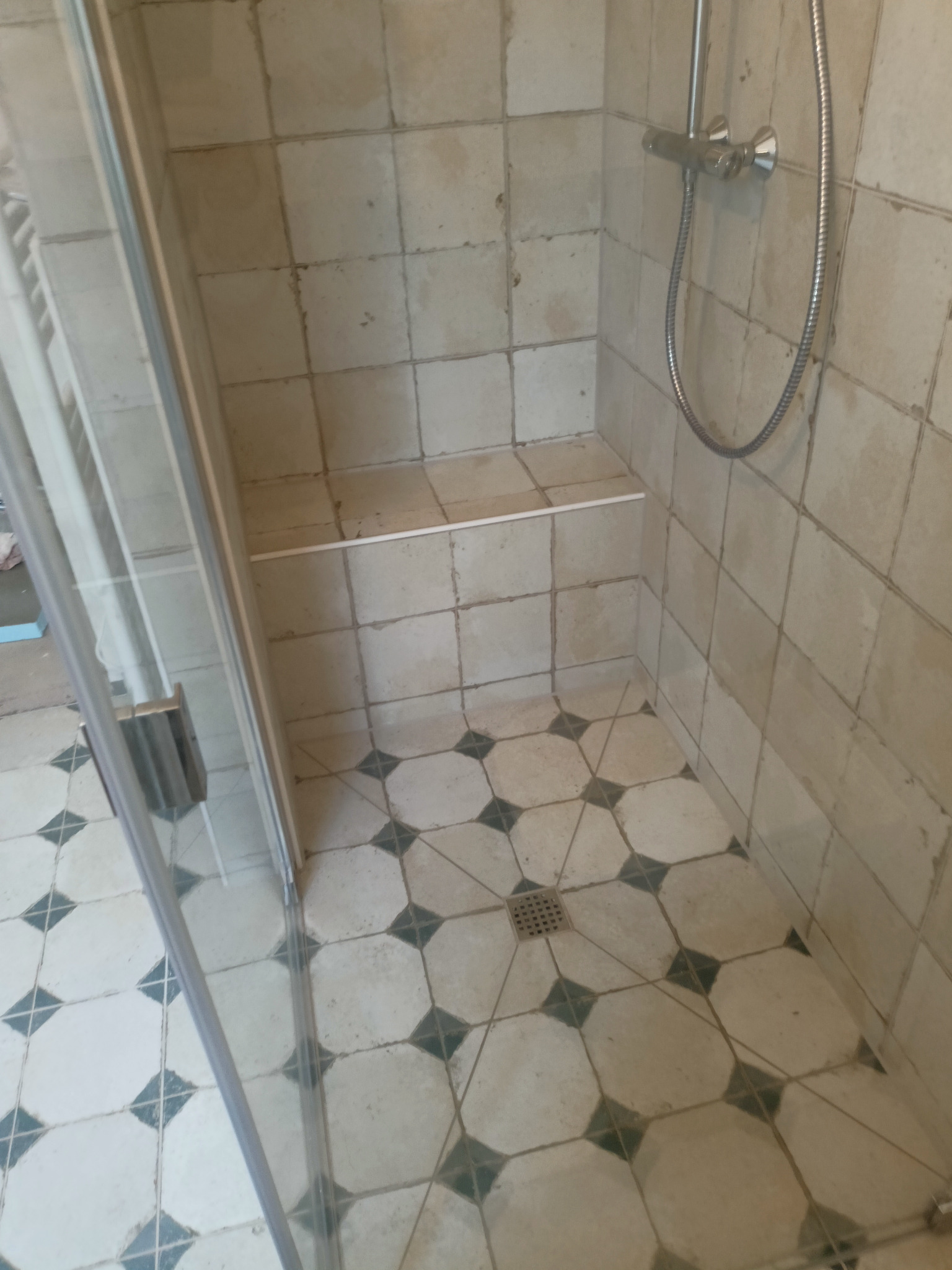 Narożna kabina prysznicowa z siedziskiem wyłożona beżowymi płytkami z ciemnymi akcentami na podłodze, widoczny odpływ liniowy i bateria prysznicowa.