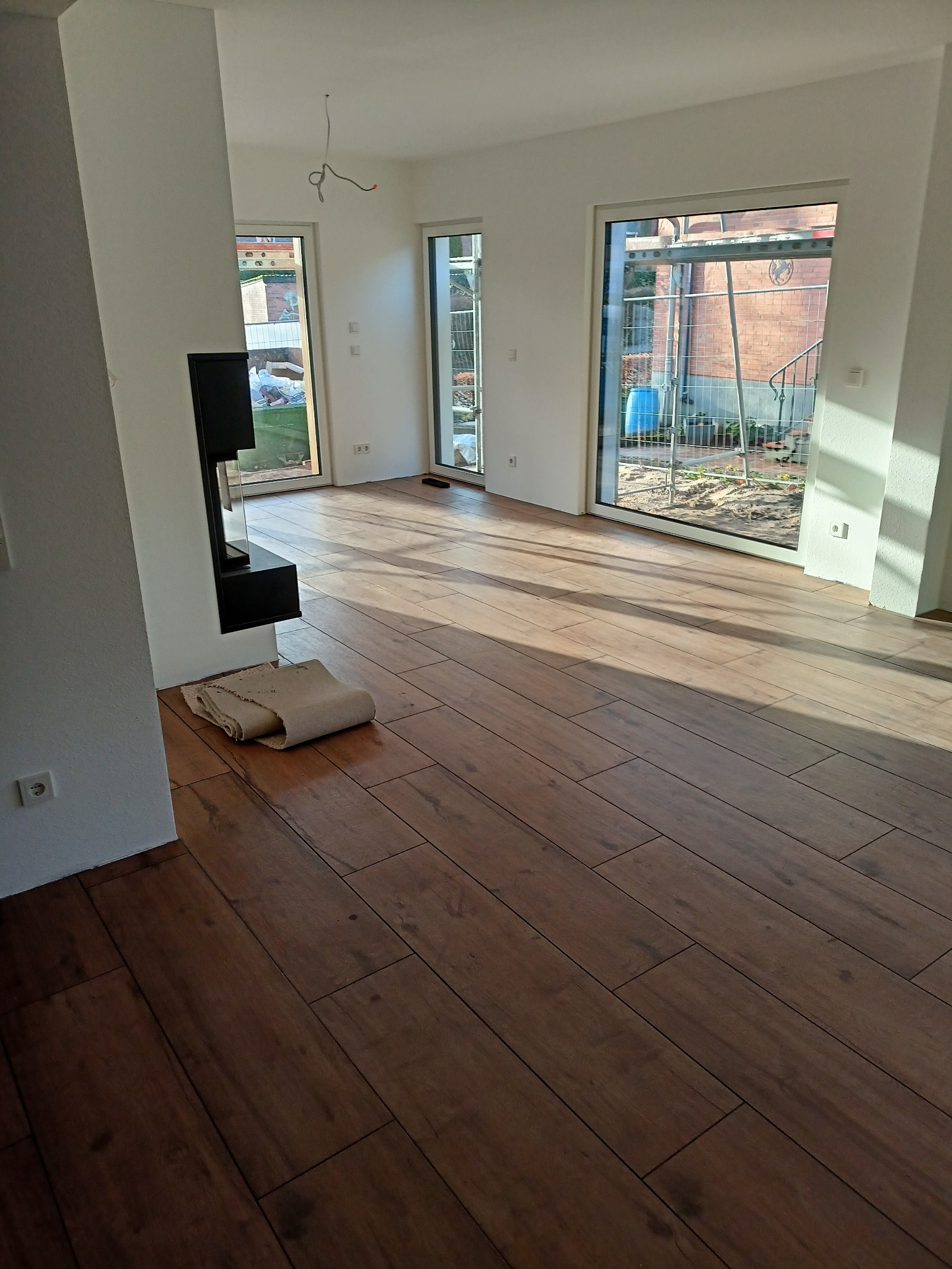 Nowo położona, ciemna, drewnopodobna podłoga w jasnym wnętrzu z dużymi oknami. Zrolowana mata ochronna leży obok czarnego kominka. Na zewnątrz widoczne rusztowania.