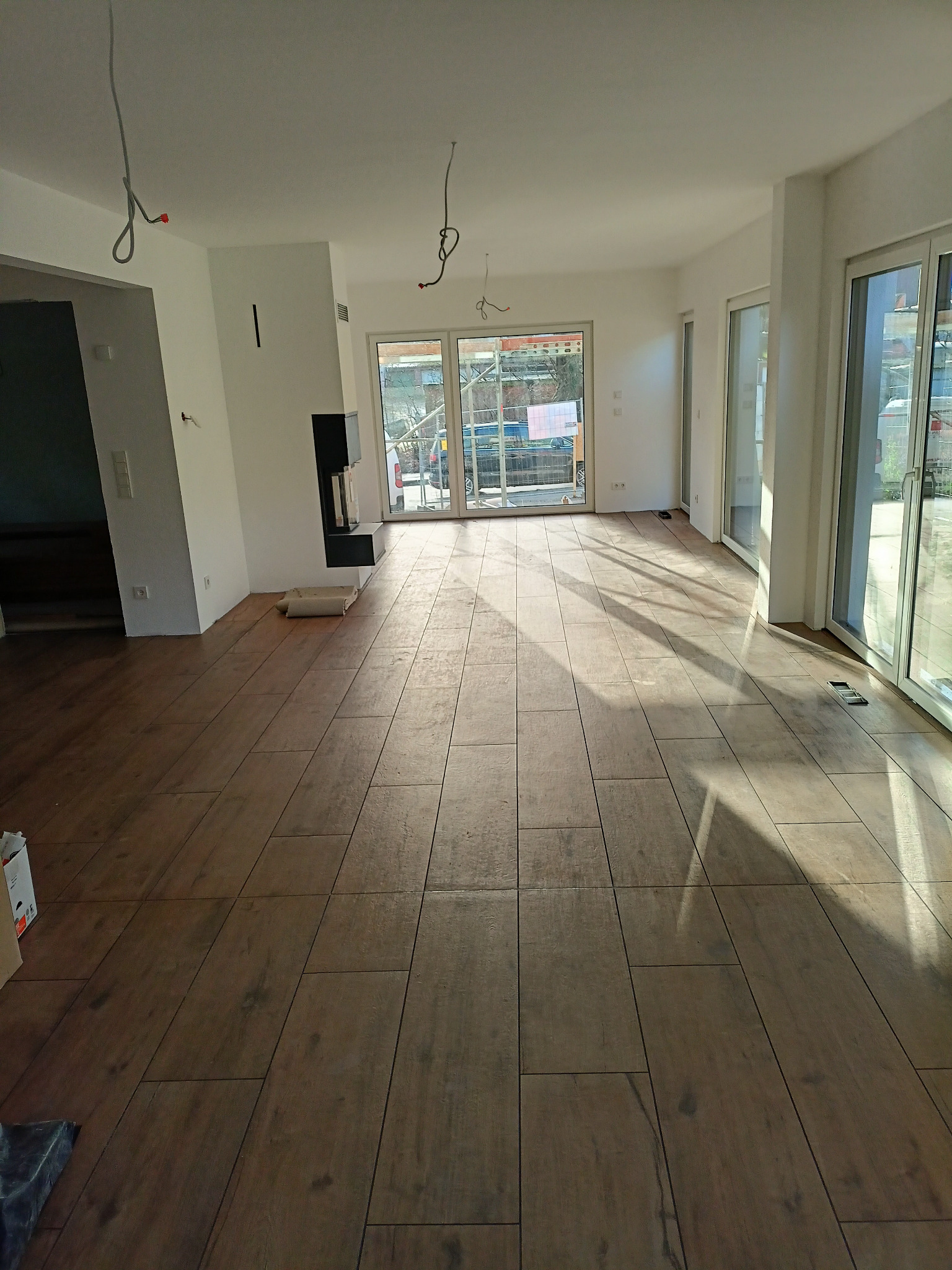 Salon po remoncie z nową, drewnopodobną podłogą z płytek, widoczne przygotowane kable do oświetlenia sufitowego i fragment kominka.