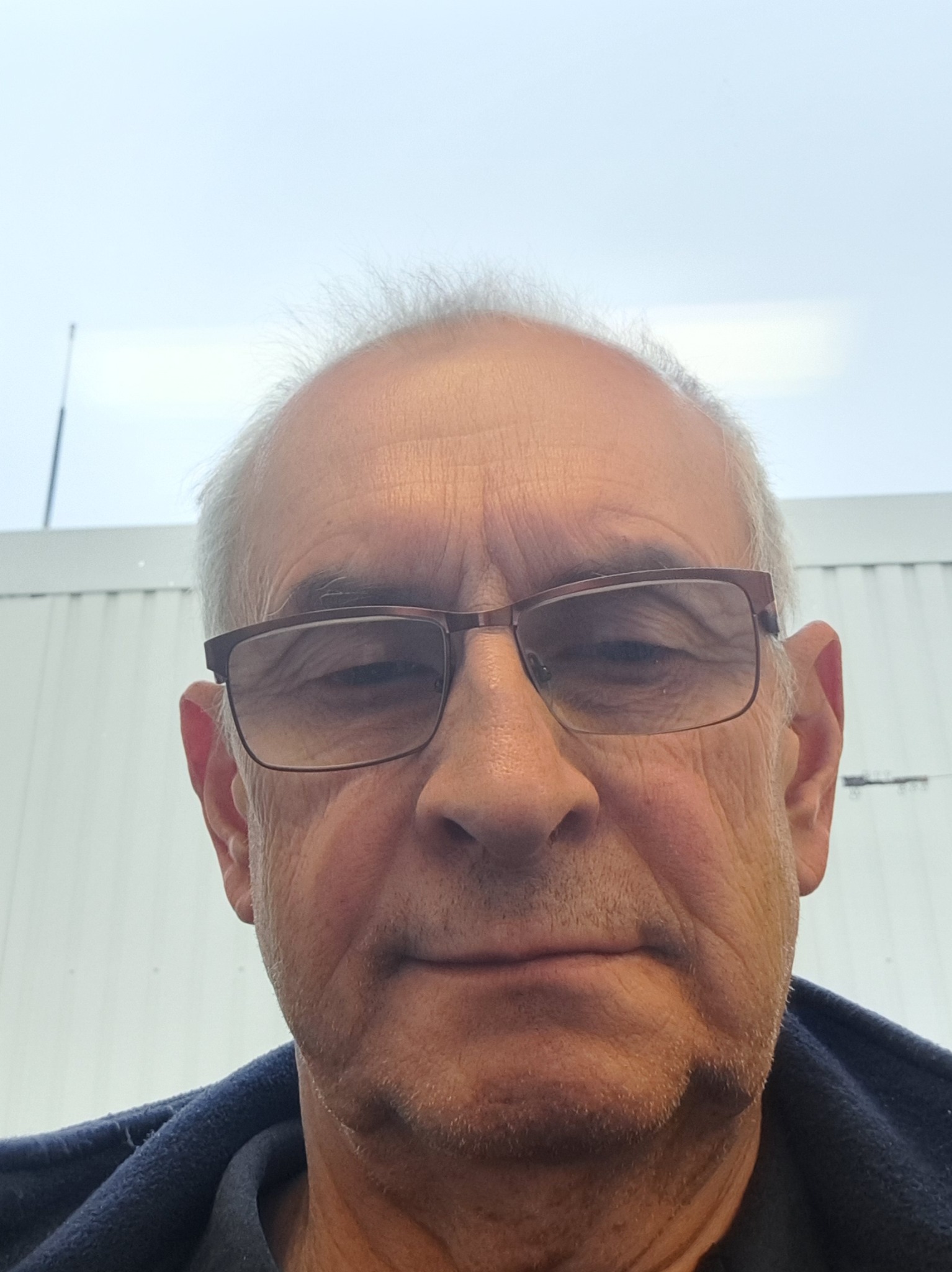 Portret starszego mężczyzny w okularach, z widocznymi zmarszczkami i siwymi włosami, sfotografowanego z dołu na tle jasnego nieba i fragmentu białej ściany.